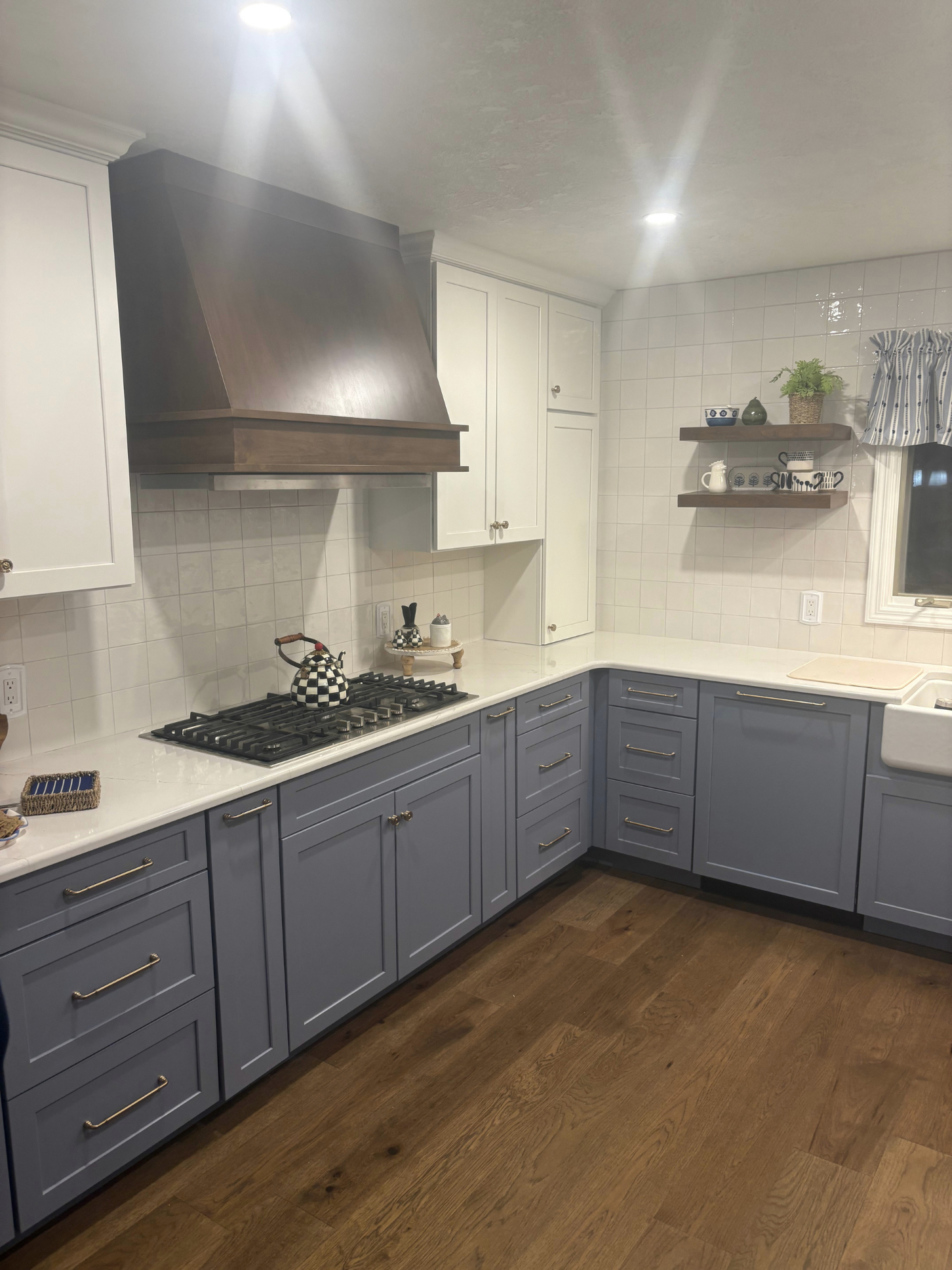 A kitchen with blue cabinetry, white countertops, a gas stove, a wooden range hood, white tiled walls, open shelves with dishes and plants, and a window with a striped curtain.