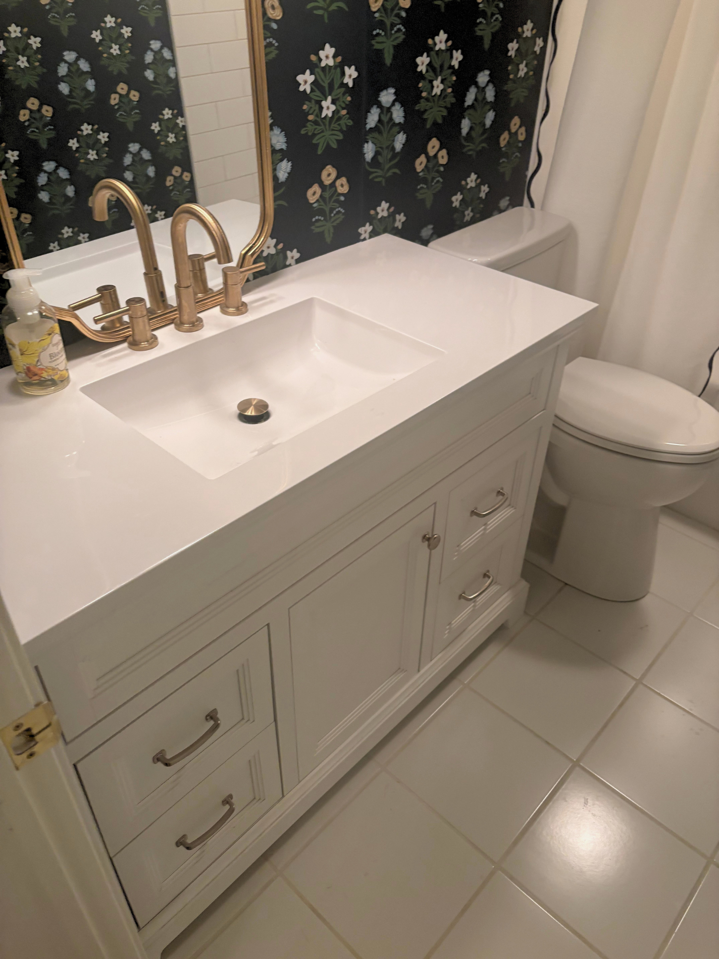 A bathroom with a white vanity and sink, gold fixtures, a mirror, and a toilet. The wall behind the sink has black floral wallpaper, and the floor is tiled in white.