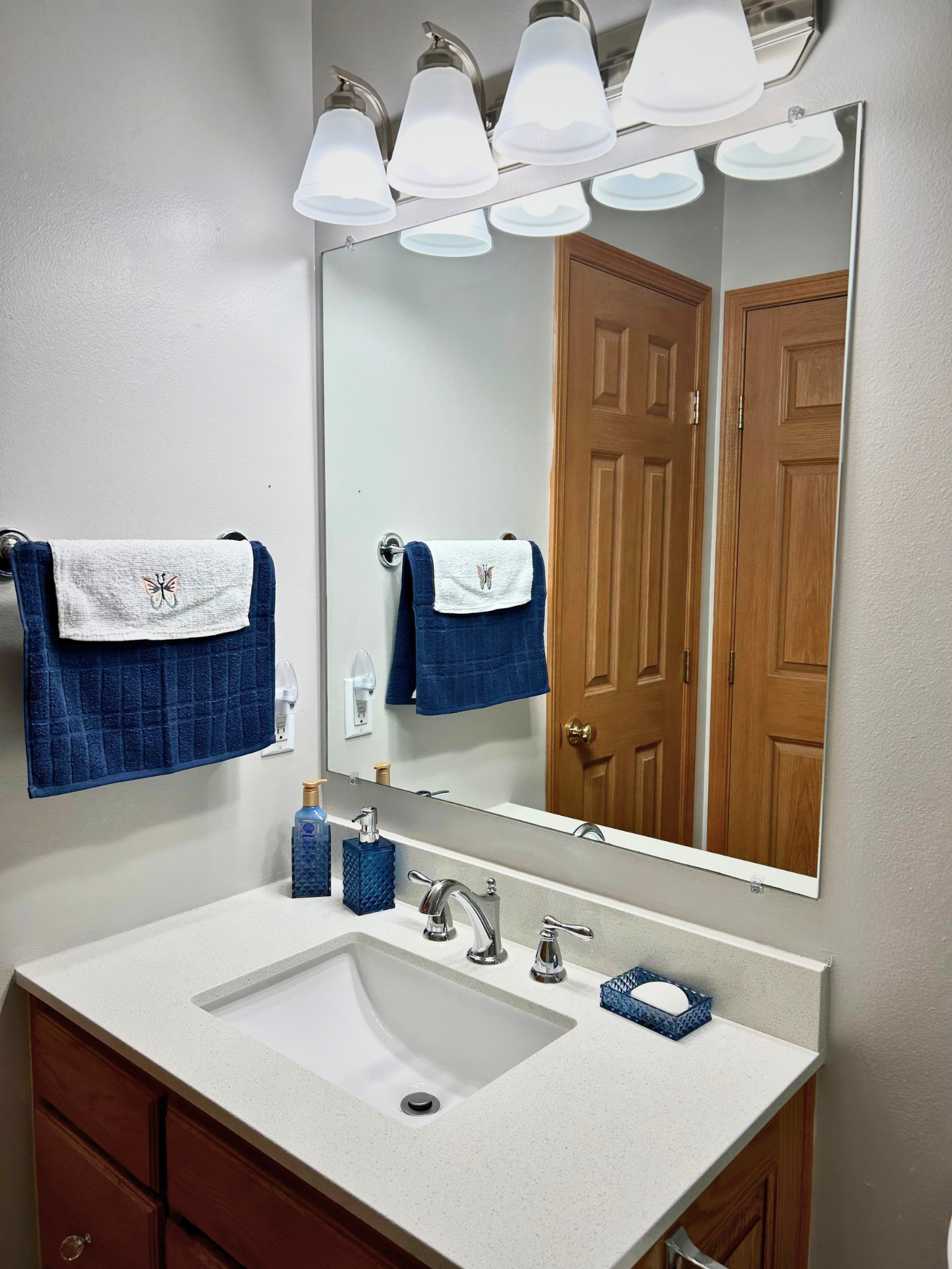 A bathroom vanity with a white countertop, a rectangular sink, a silver faucet, and matching soap dispenser and tissue box in blue. Above the sink is a large rectangular mirror. On the left side, two hand towels with butterfly embroidery hang on a to