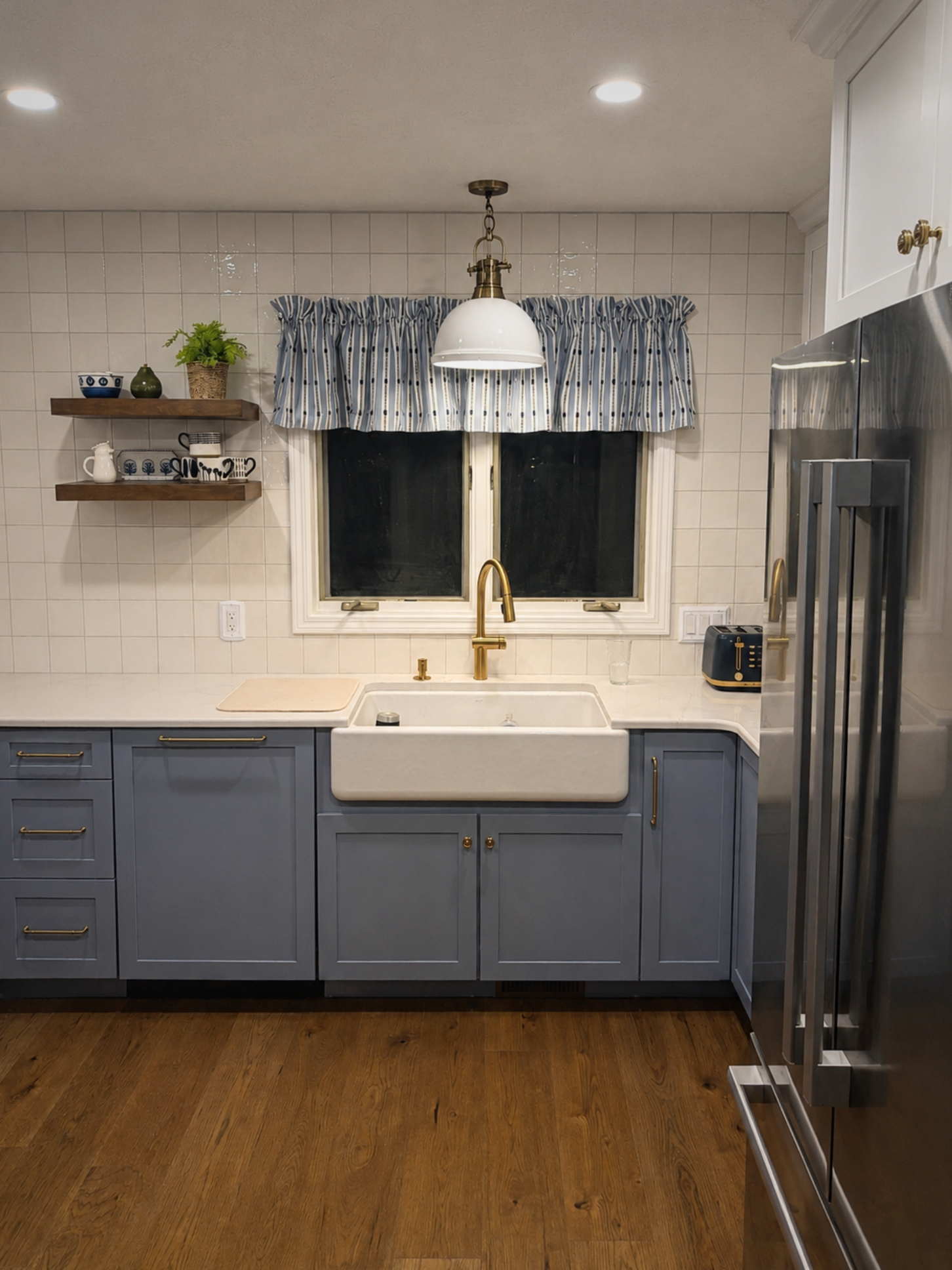 Modern kitchen with blue cabinets, white tile walls, a white farmhouse sink with gold fixtures, wooden open shelves with dishes and a potted plant, window with blue and white striped curtains, and stainless steel appliances, all on a wooden floor.