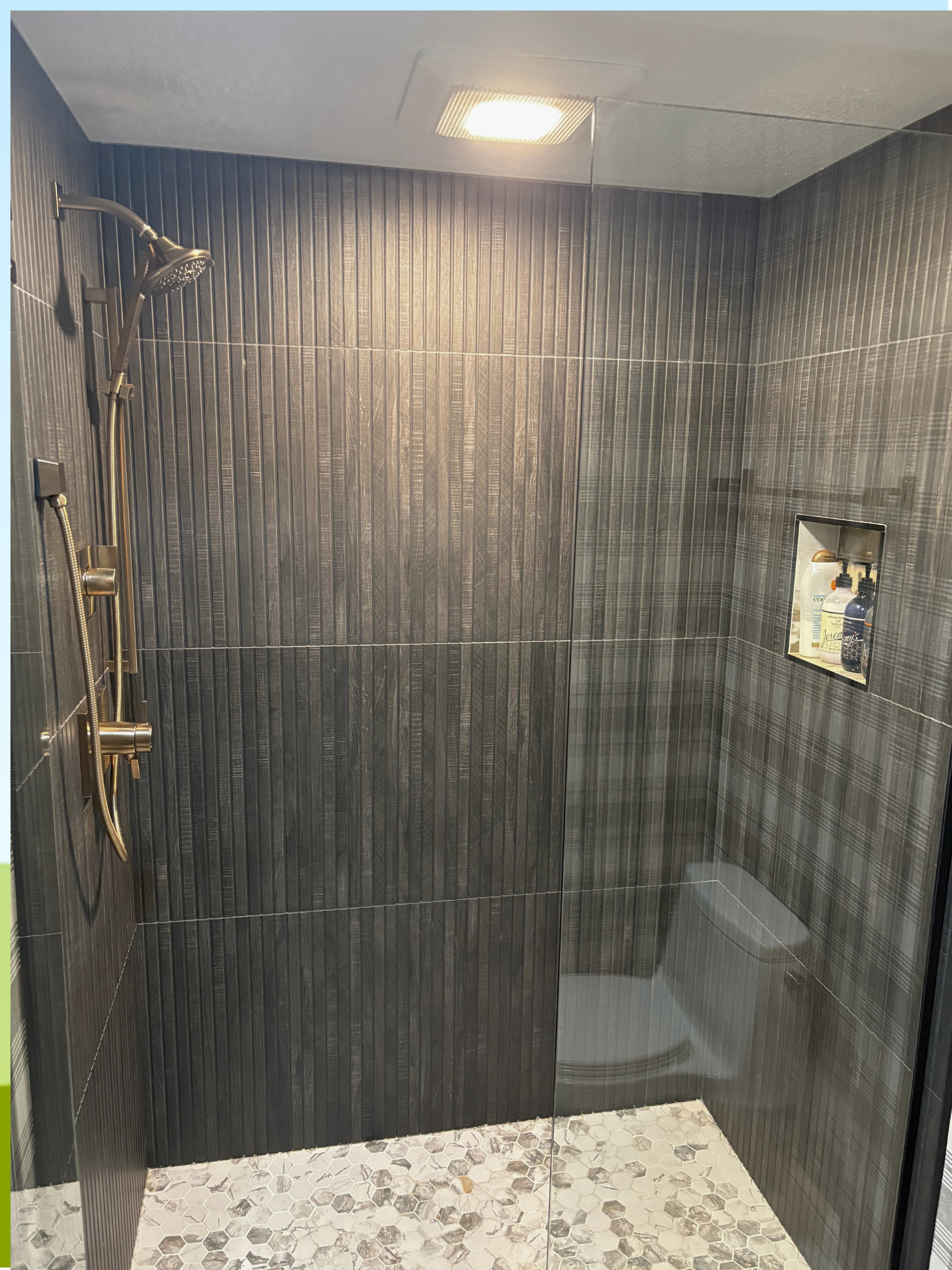 A modern walk-in shower with dark vertical tiled walls, a ceiling-mounted shower head, and a small built-in wall niche holding bottles of shampoo and body wash. The shower has a glass door and a floor with hexagonal tiles.