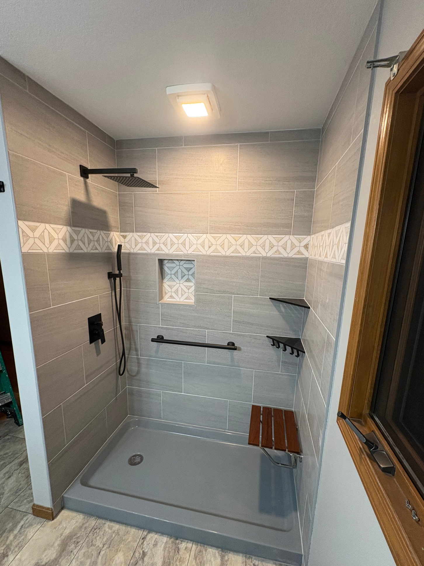 Modern shower with beige and gray tiles, black fixtures, a wooden shower seat, and a small built-in shelf.