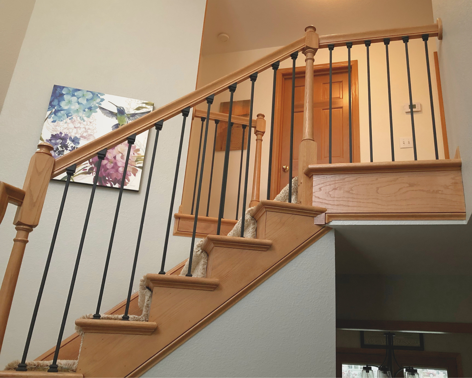 Interior view of a staircase with wooden steps and black metal spindles, and a partial view of a door at the top of the stairs. A colorful painting of a hummingbird and flowers is on the wall to the left.