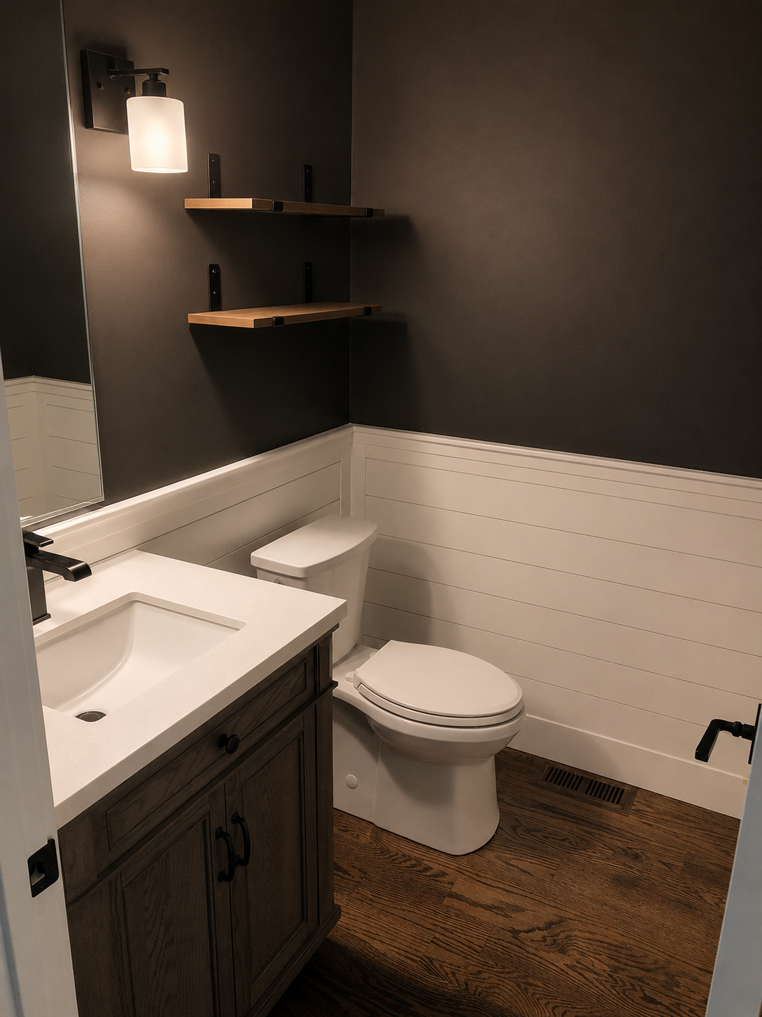 A small bathroom featuring a white sink vanity with black fixtures, a white toilet, dark wall paint, two wooden floating shelves, and a wall-mounted light fixture.
