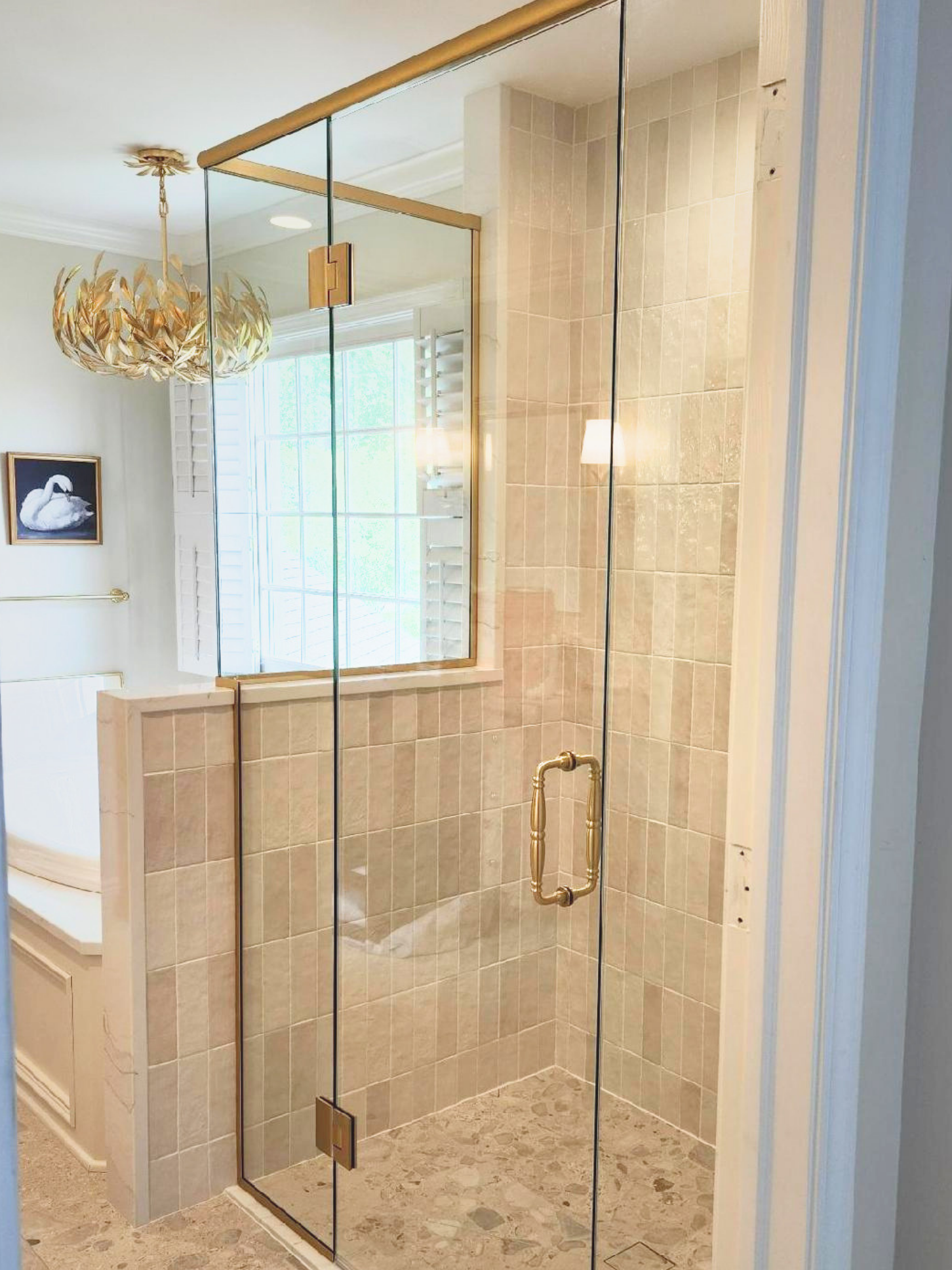 A glass shower enclosure with gold hardware in a bathroom with beige tiled walls and a window with white shutters.