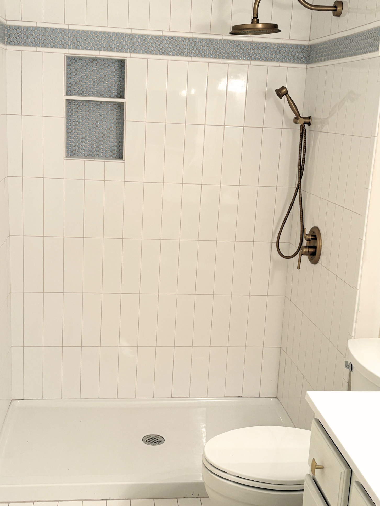 A bathroom with white tiled walls, a walk-in shower with a bronze showerhead and a handheld shower wand, a small built-in shelf with blue mosaic tiles, a white toilet with a closed lid, and a white vanity cabinet with a single drawer, a beige handle,