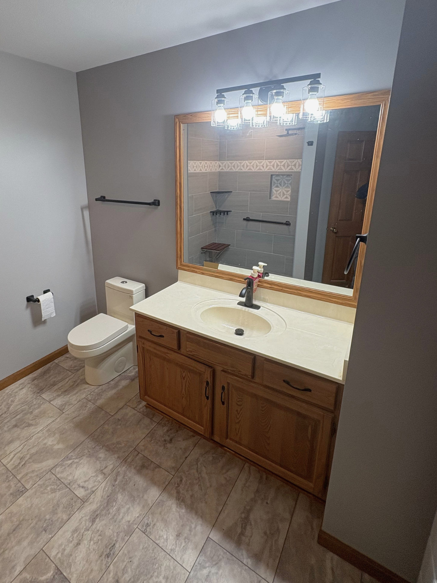 Bathroom with a wooden vanity, a mirror, a light fixture, a toilet, and a tiled shower area with a wooden bench and black towel bars.
