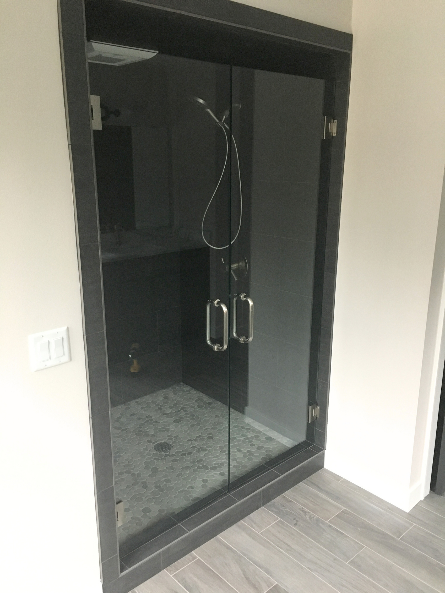A glass shower enclosure with black tiles, metal handles, and a grey stone tile floor, located next to a white wall with a light switch.