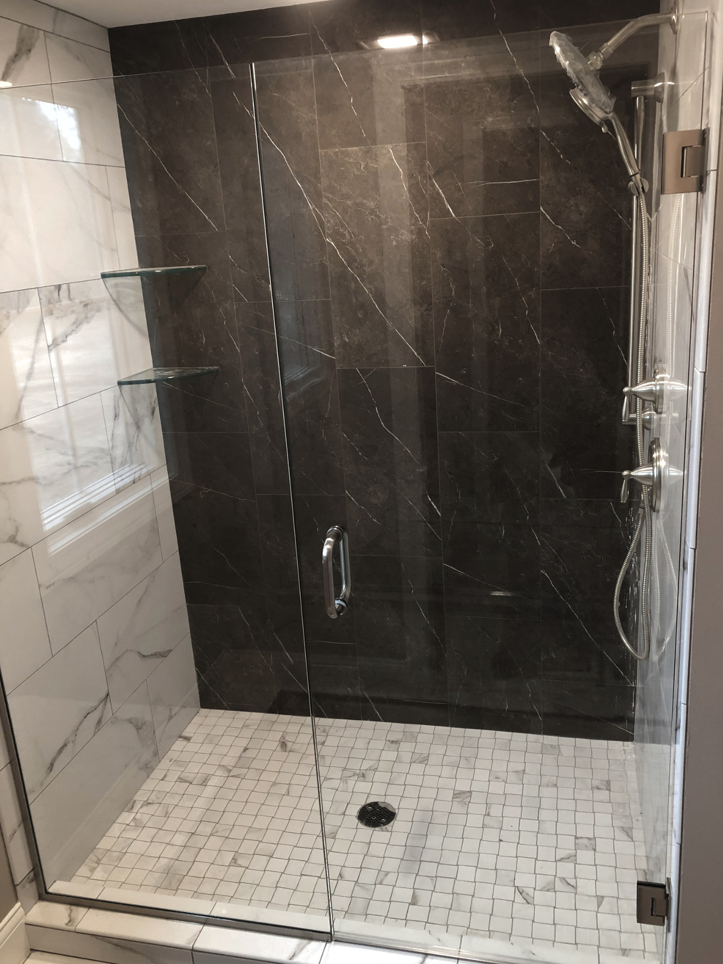 A shower with glass door, black marble tiles on the back wall, white marble tiles on the left wall, and a white tiled shower floor with a circular drain. There are two glass corner shelves on the left wall, and a silver showerhead with a flexible hos