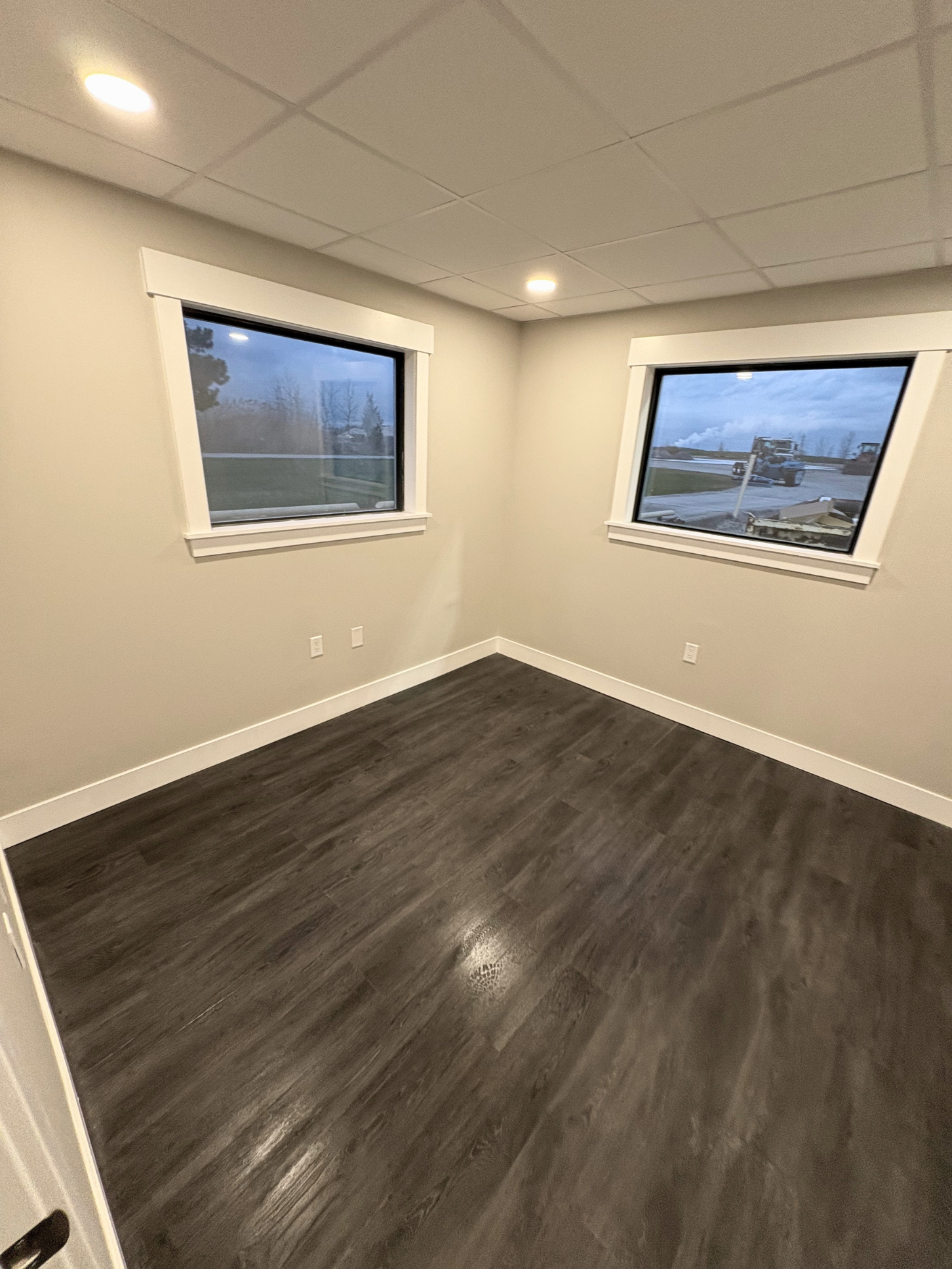 Empty room with two large windows, dark wood flooring, white walls, and ceiling with recessed lighting.