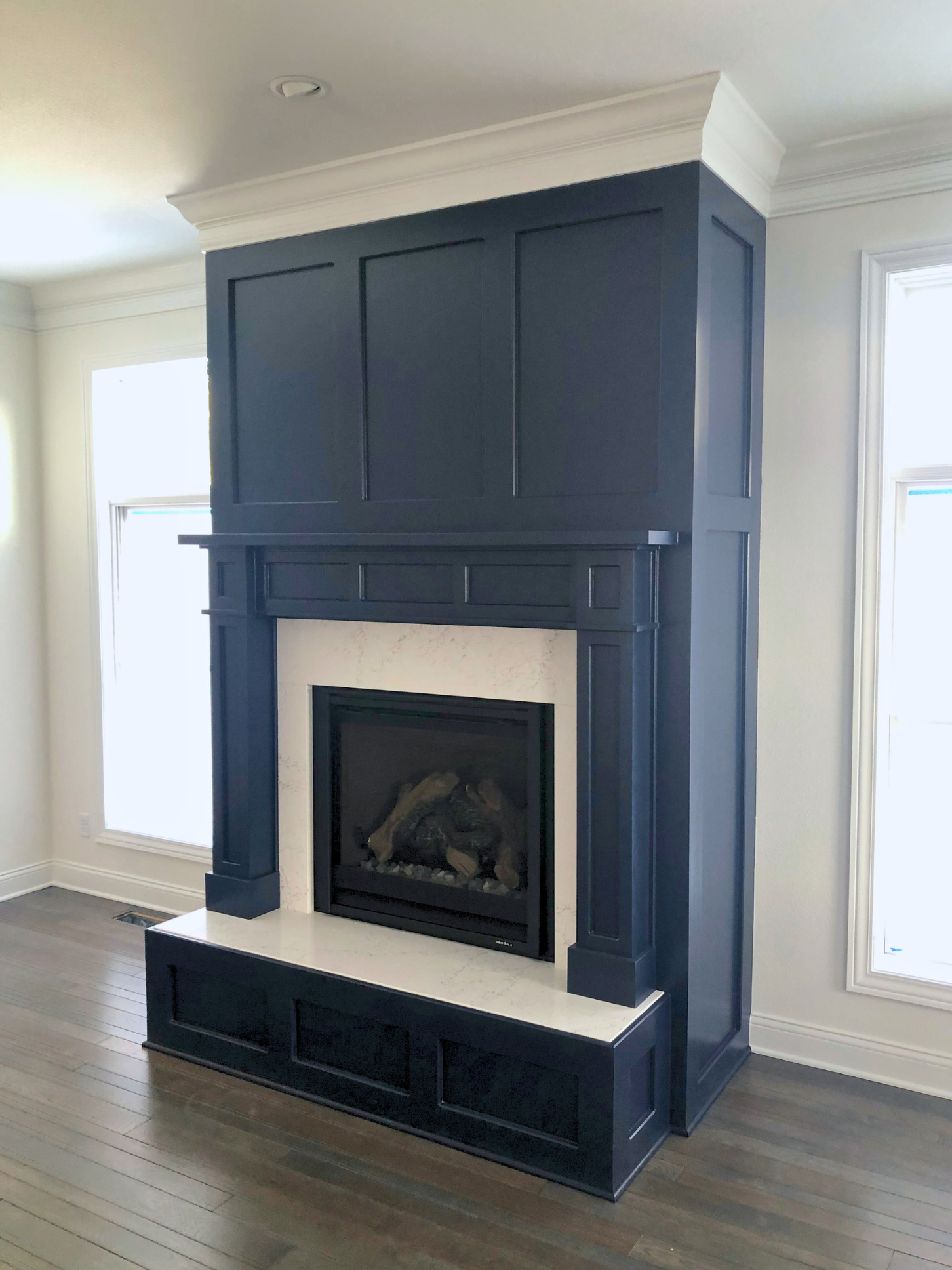 Interior view of a living room corner showing a dark navy blue built-in fireplace mantel with white marble surround, flanked by two large windows with white trims, and hardwood flooring.