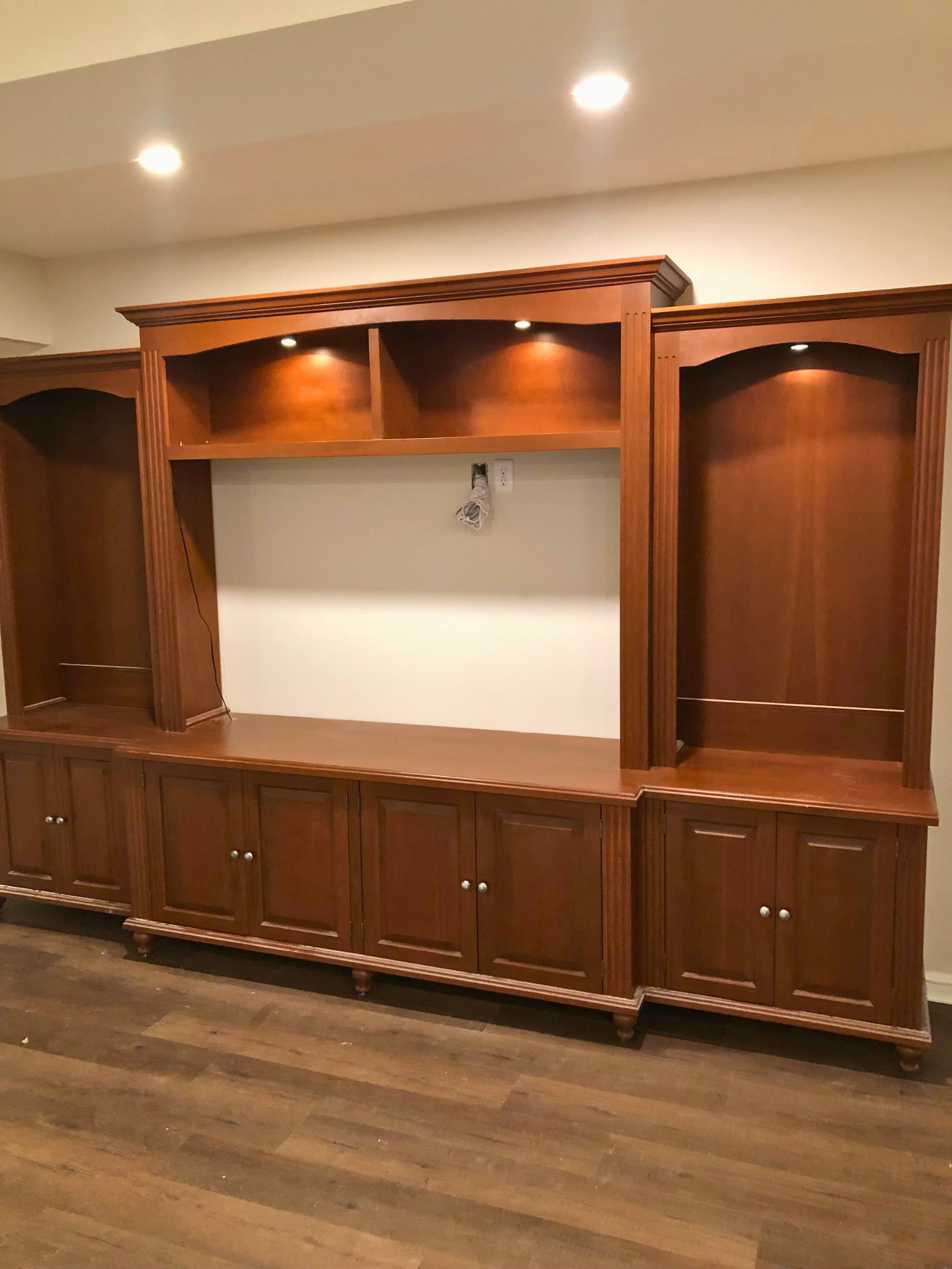 Empty wooden built-in wall unit with cabinets and open shelves, some spotlights above, on a light-colored wall, with hardwood flooring.