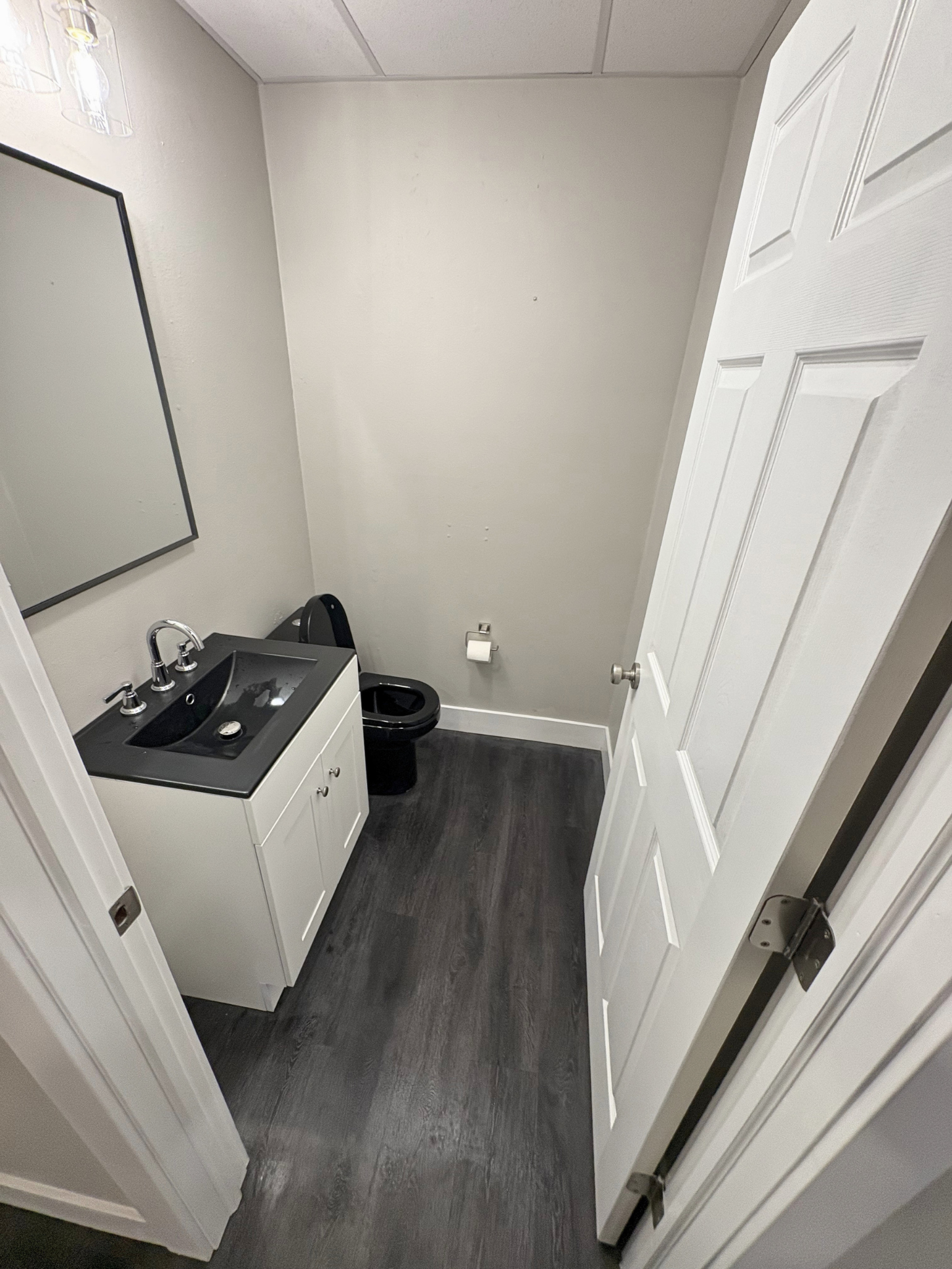 A small bathroom with a white vanity and black sink, a black toilet, a mirror, a wall-mounted cabinet, and a toilet paper holder. The floor is dark wood or laminate, and the walls and door are white.
