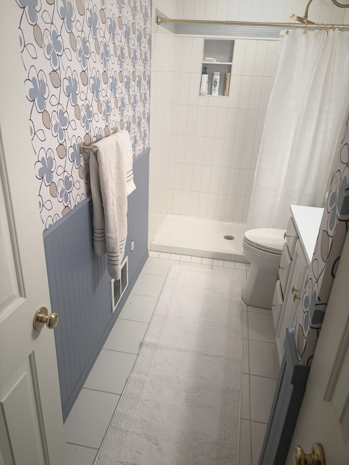 A bathroom with a patterned wallpaper on the left, a white shower with a curtain on the right, a white toilet, and a white cabinet. There is a white bath mat on the tiled floor.