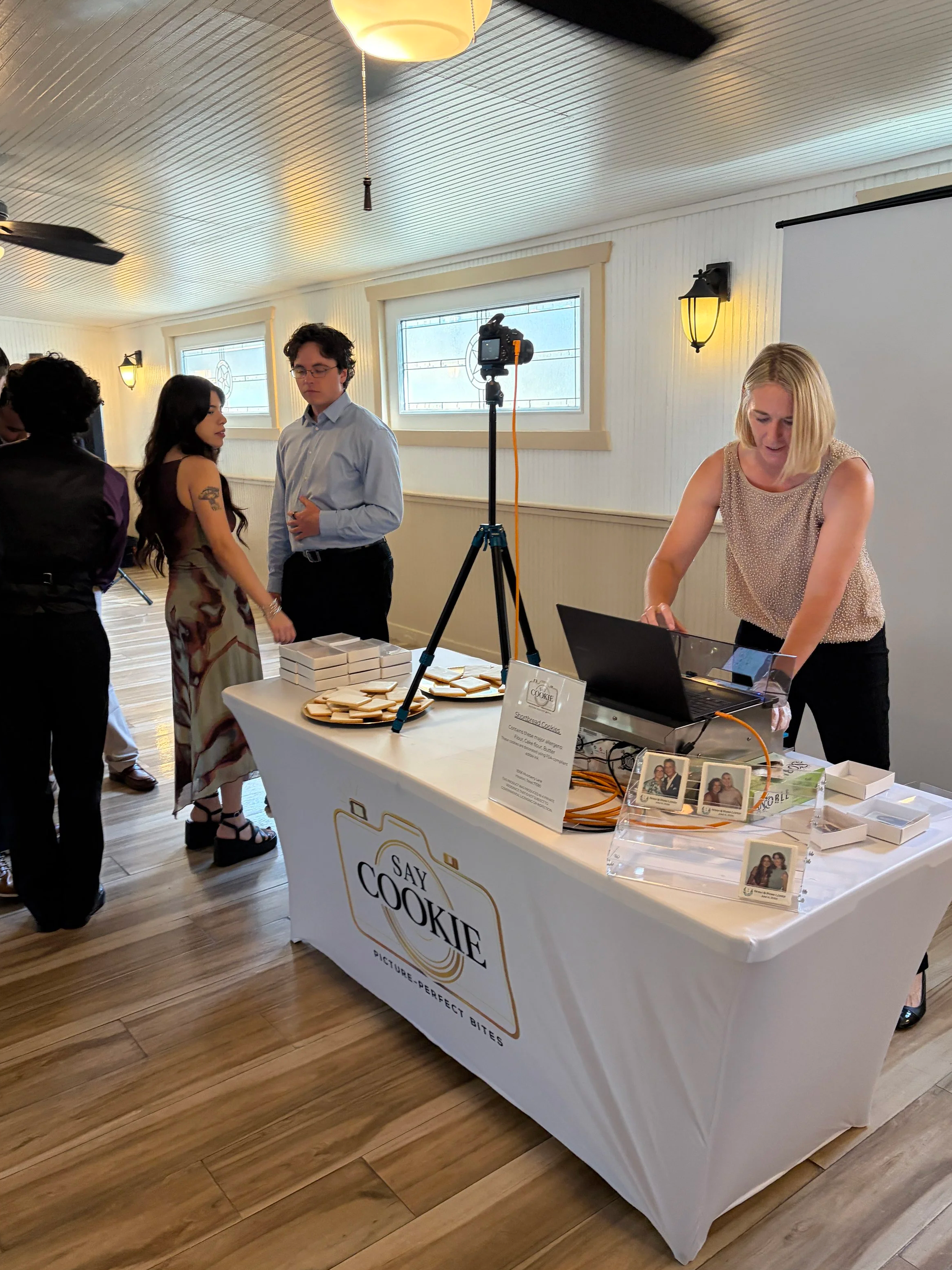 Several people standing around a table with cookies at a social event or gathering indoors. One woman at the table is working on her laptop, and there is a sign on the table that says "Say Cookie." The room has white walls with windows, mounted lights, and ceiling fans.
