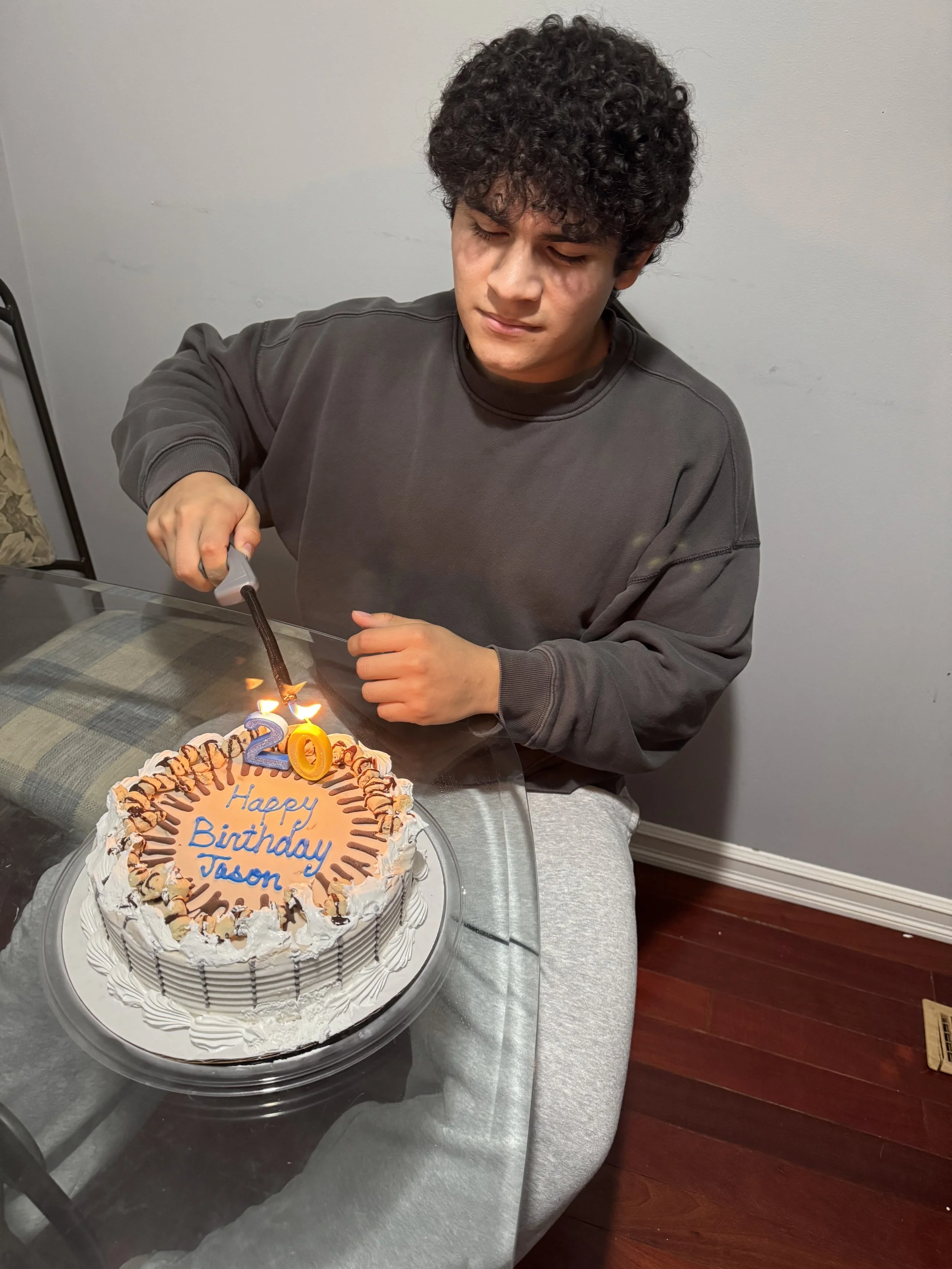 A young man with curly hair celebrating a birthday, lighting candles on a birthday cake that has a message reading "Happy Birthday Jason" with a "20" candle on top, sitting at a table with a gray tablecloth against a plain wall.