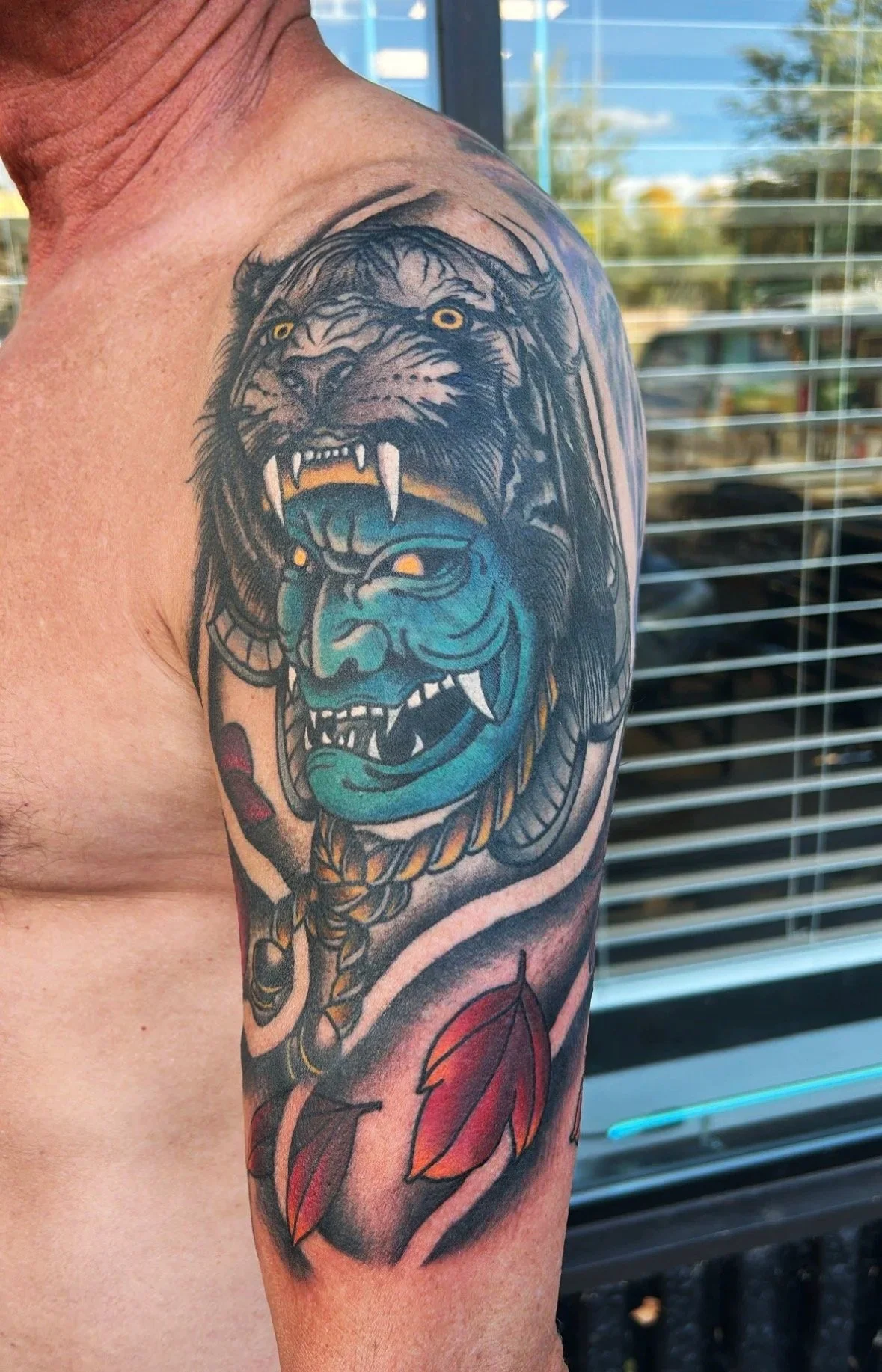 Japanese arm tattoo featuring a fierce lion headdress over a detailed blue Japanese style face, with falling red leaves underneath.