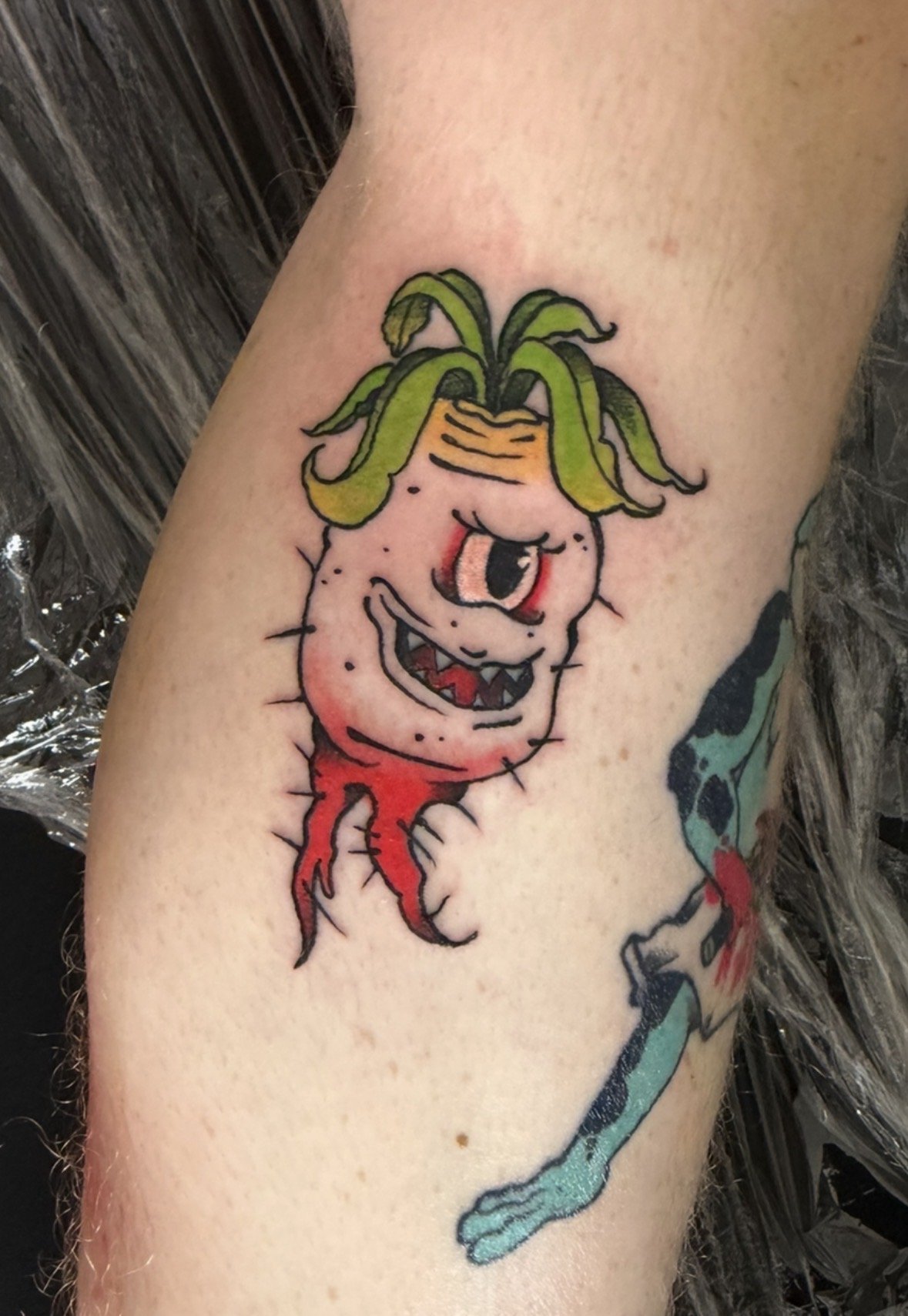 Colorful tattoo of a mischievous cartoon-style plant creature with a wide, toothy grin, one eye, green hair, pink skin with black spots, red roots for legs, on a person's arm.