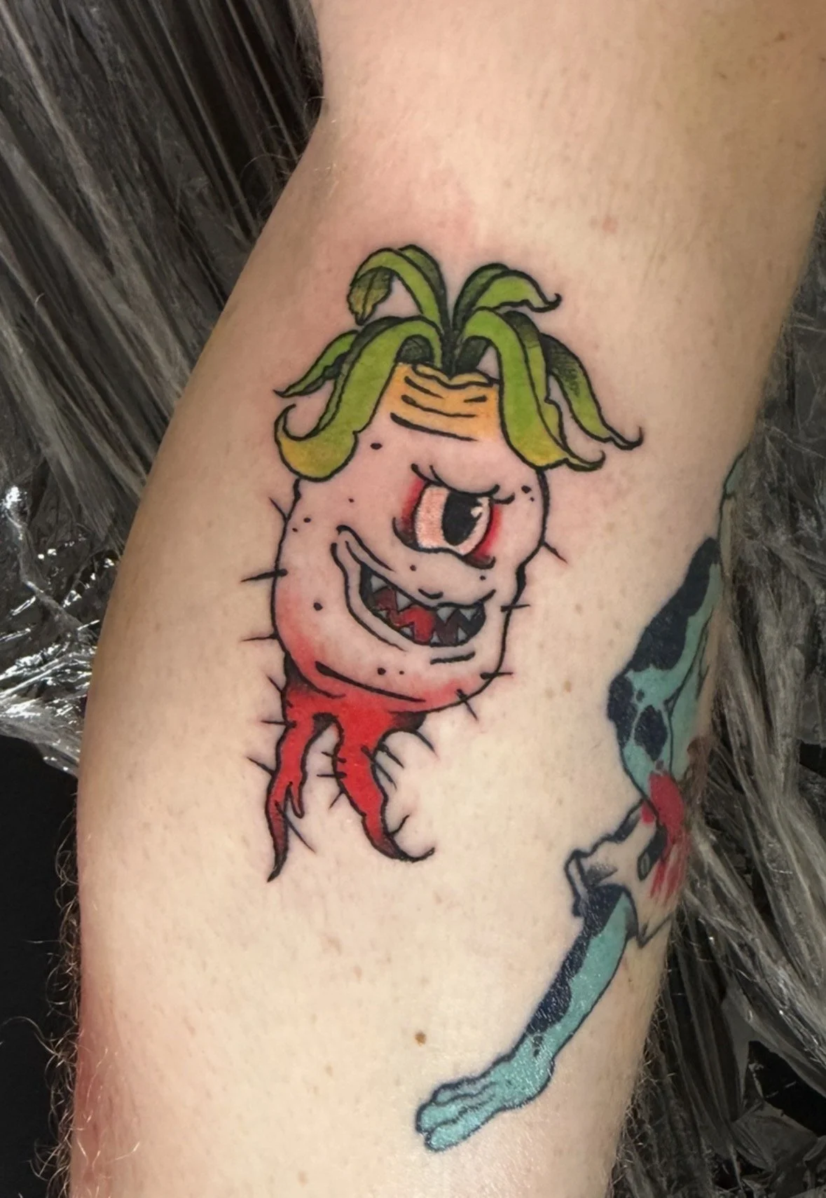 Colorful tattoo of a mischievous cartoon-style plant creature with a wide, toothy grin, one eye, green hair, pink skin with black spots, red roots for legs, on a person's arm.