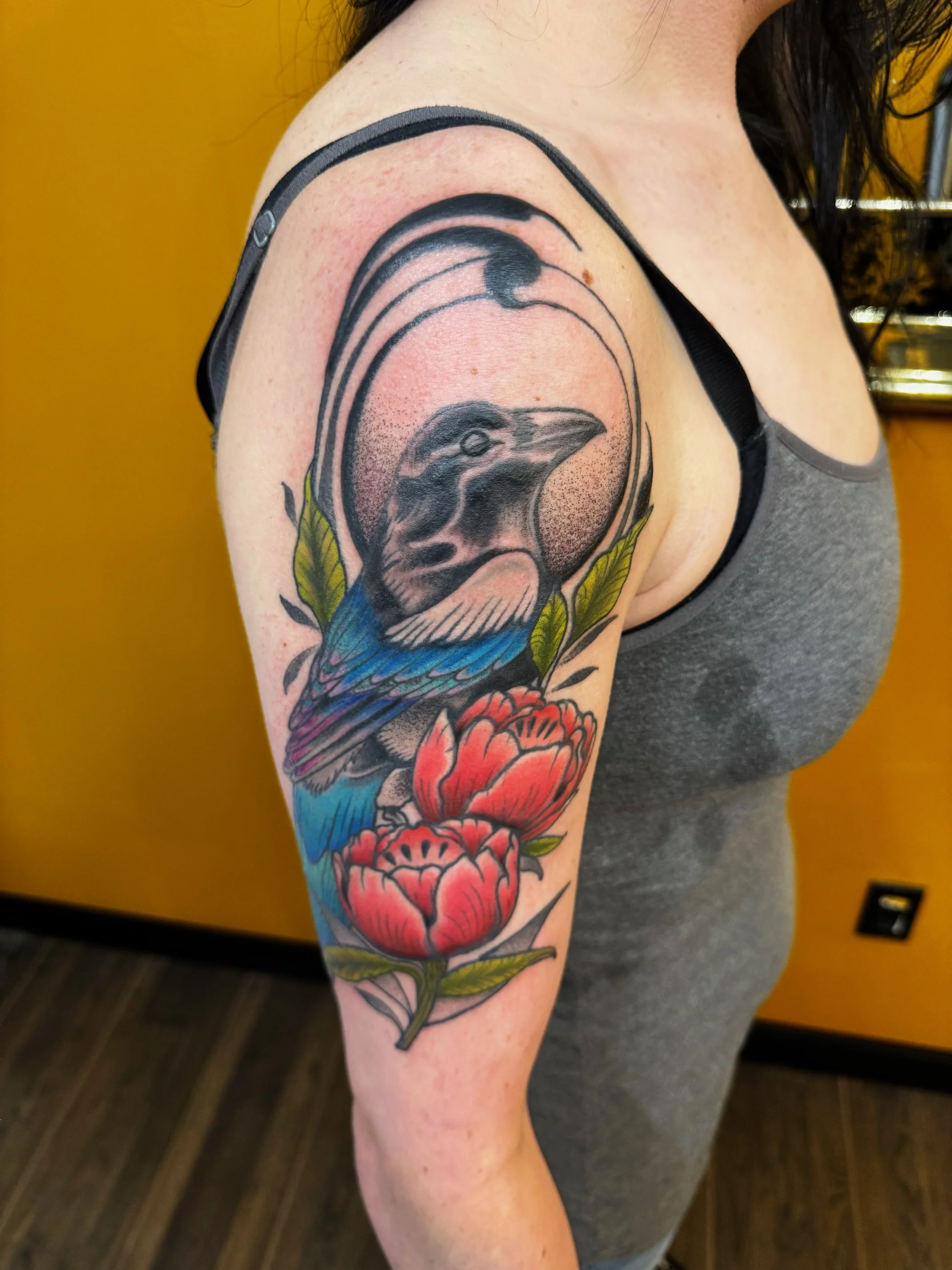 Tattoo of a crow with blue and purple touches on its feathers, with red and green floral and geometric elements surrounding it, located on a person's upper arm.