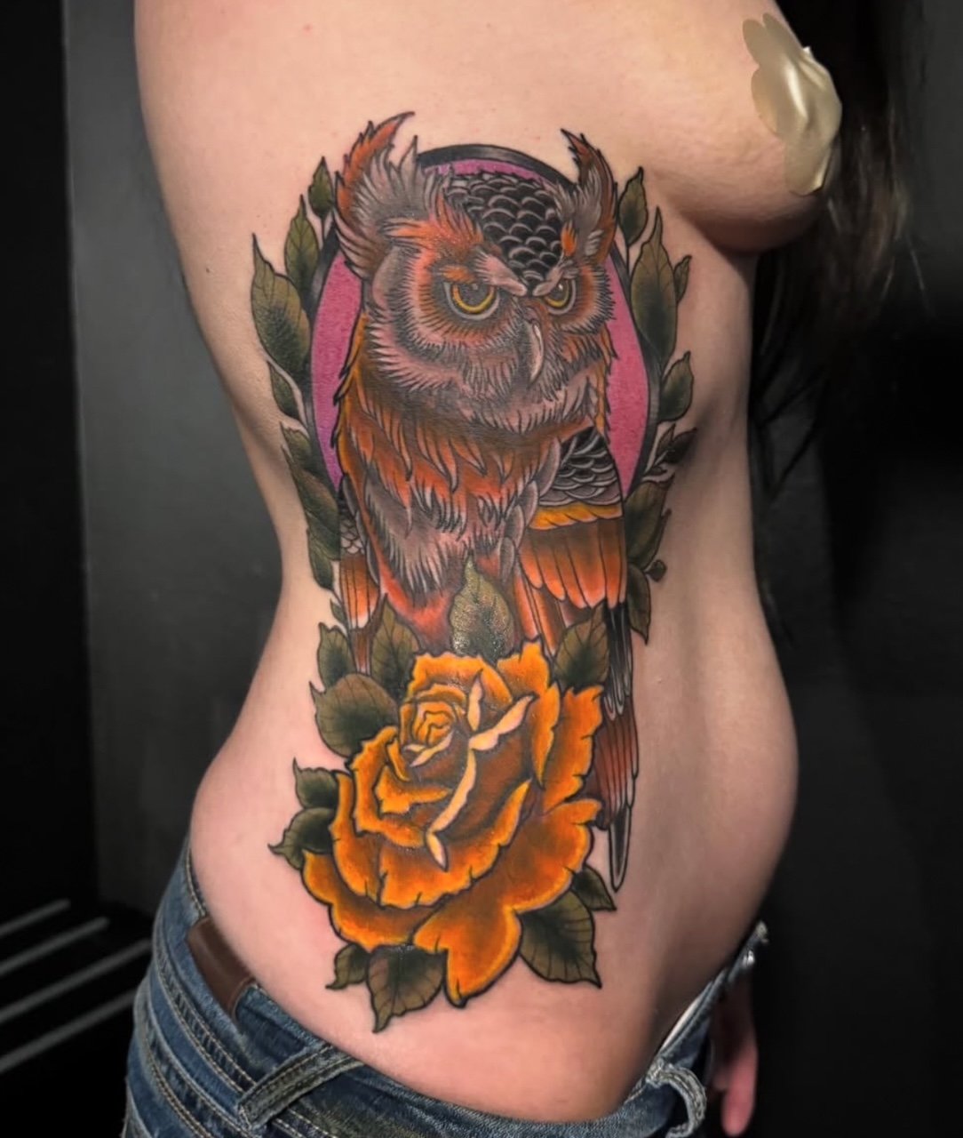 Colorful tattoo of an owl with detailed feathers, surrounded by leaves and a large orange rose on a person's side ribcage.
