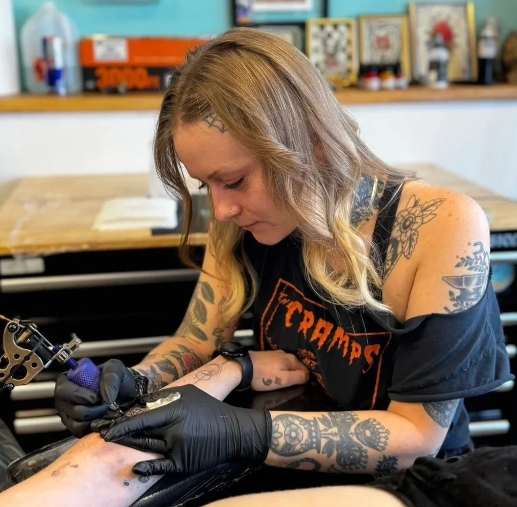 A woman with tattoos on her arms and shoulder is tattooing a person's leg in a tattoo parlor in Denver.