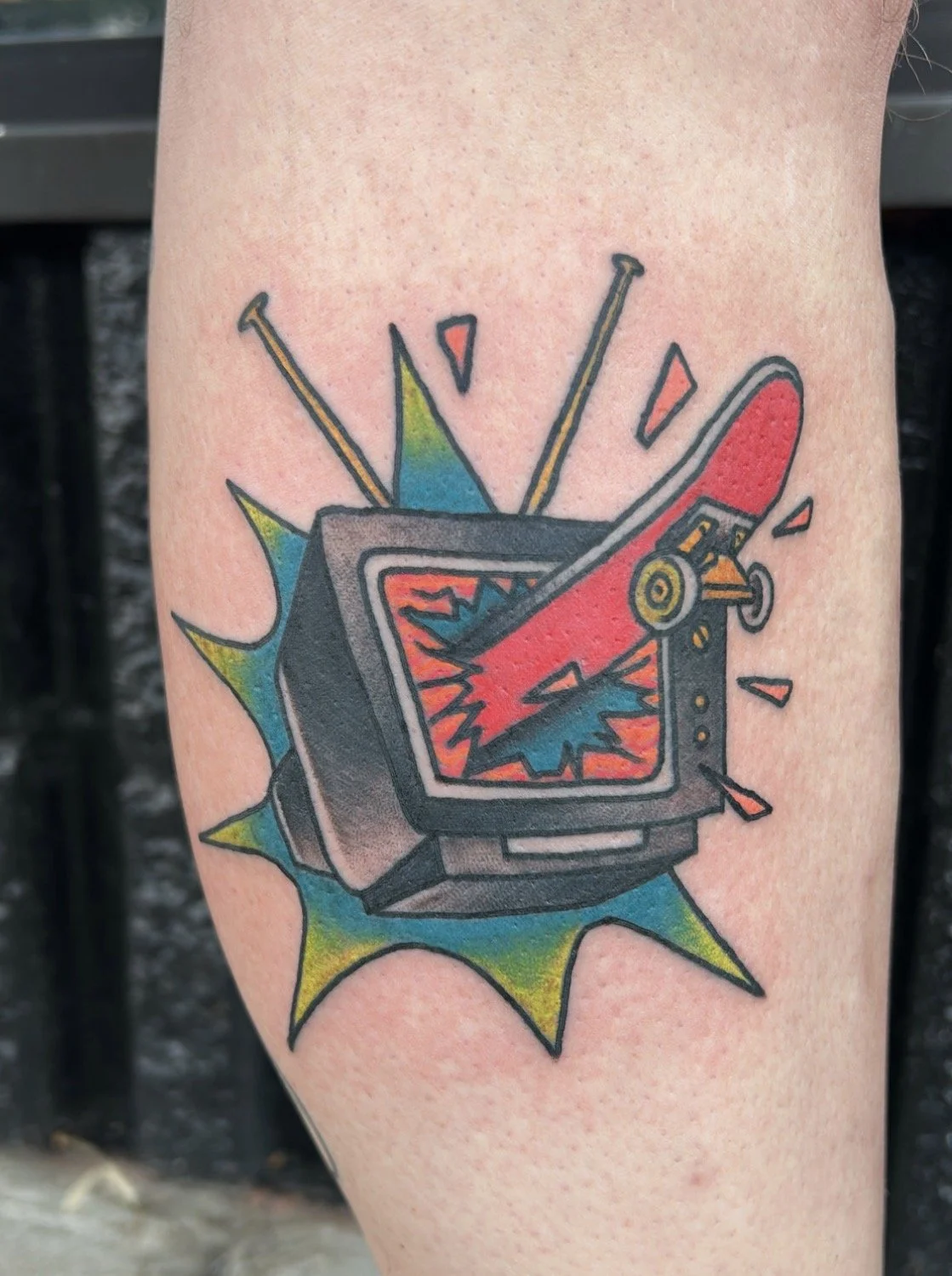 Custom tattoo of a red skateboard breaking through the screen of an old T.V. causing an explosion, with bursts around it.