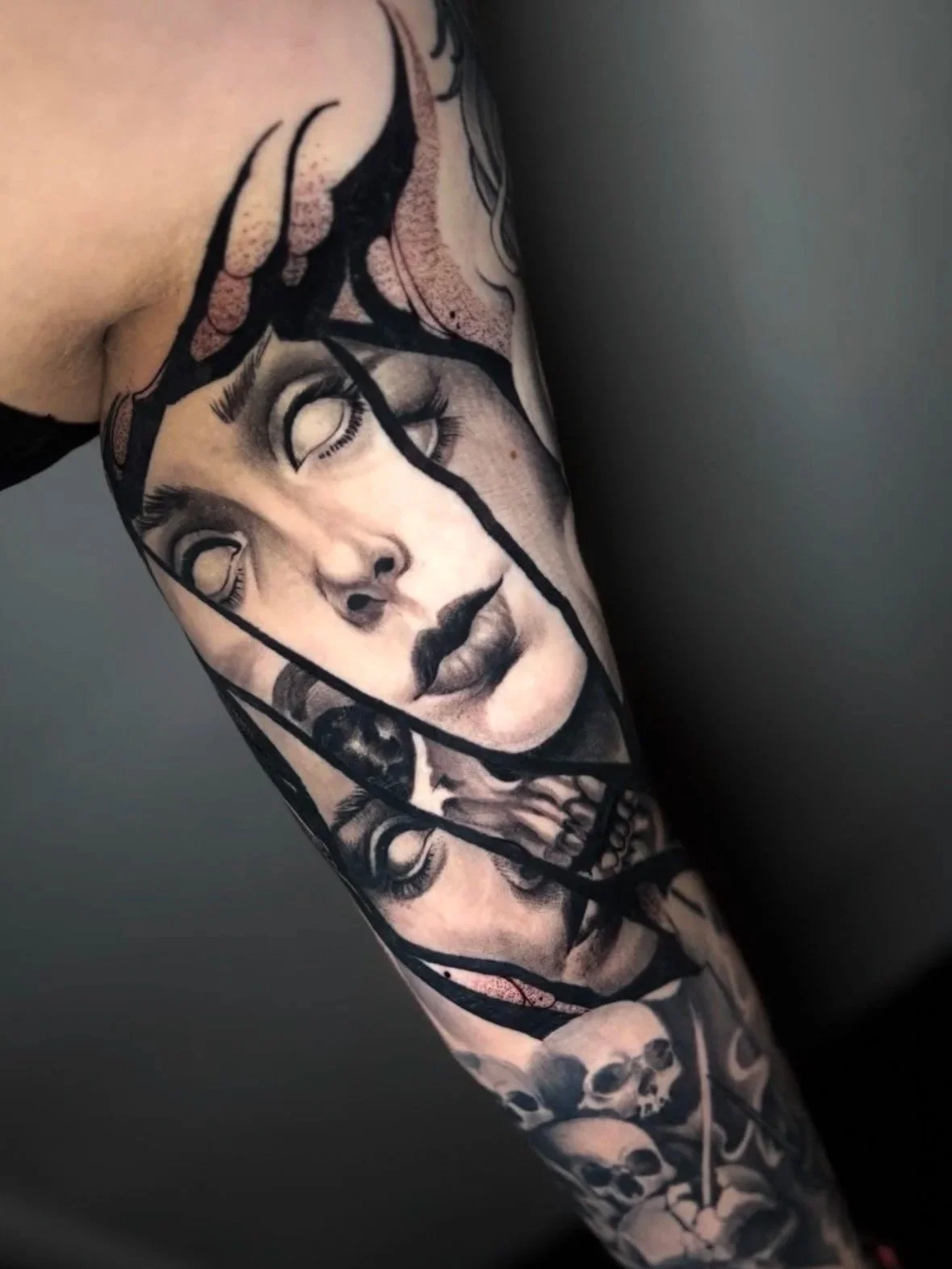Close-up of a tattoo sleeve featuring a realistic portrait of a woman with white eyes, skulls, and abstract elements surrounded by thick line work, in black and gray ink.