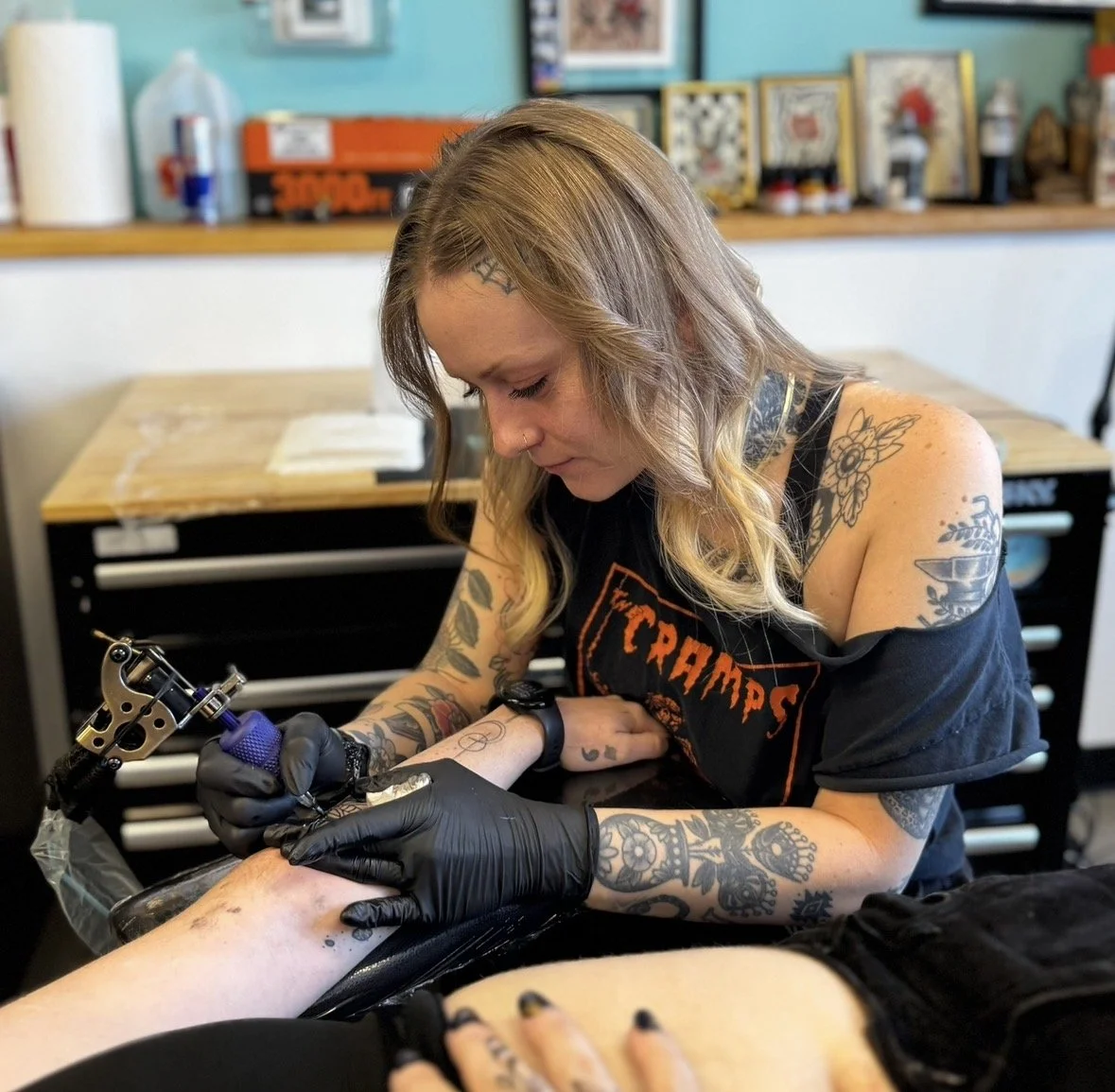 A woman with tattoos getting a tattoo on someone's leg using a tattoo machine in a tattoo studio.