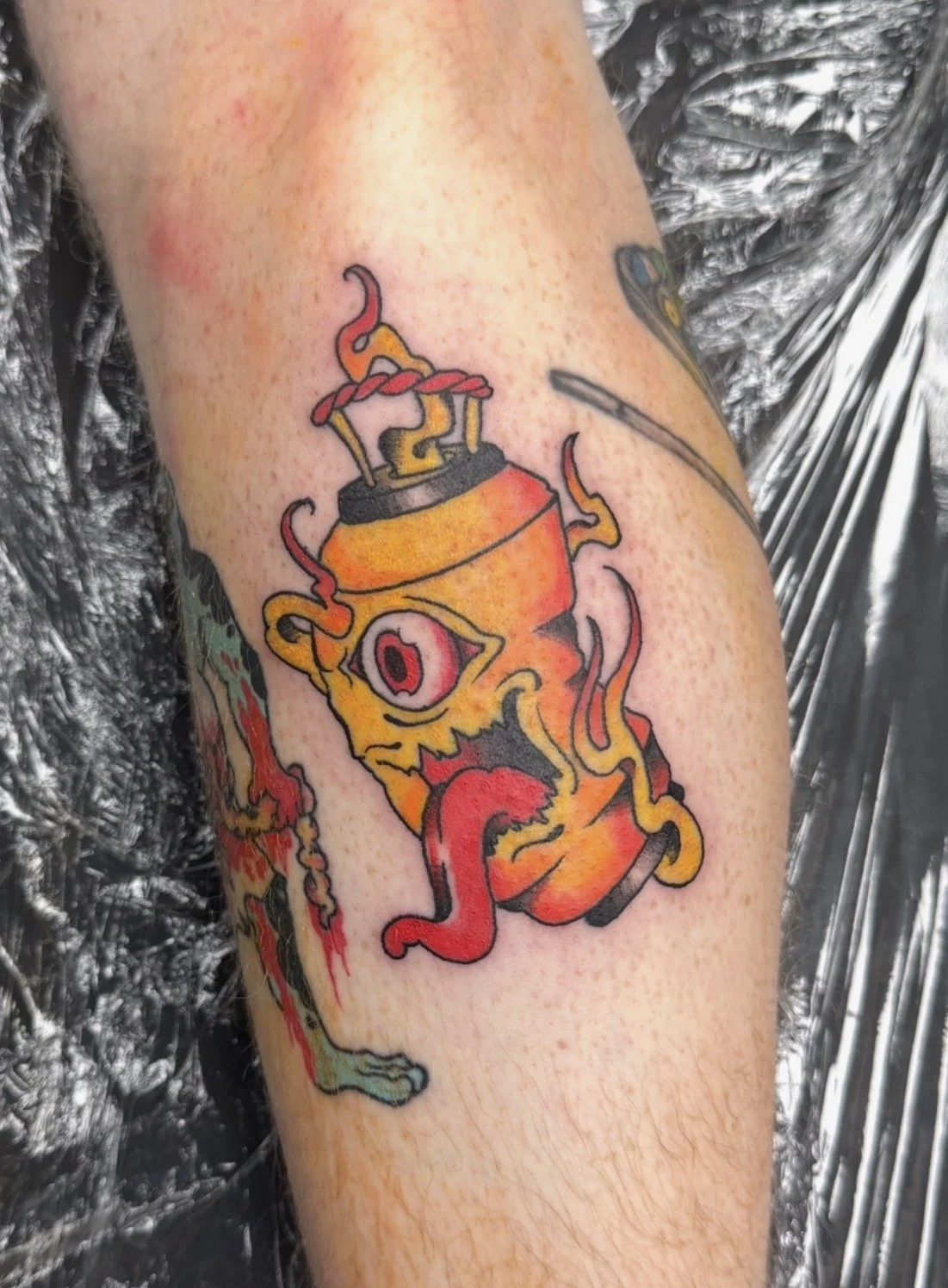 Colorful tattoo of an abstract creature with a single eye and a long tongue, featuring orange, red, yellow, and black colors, on person's arm.
