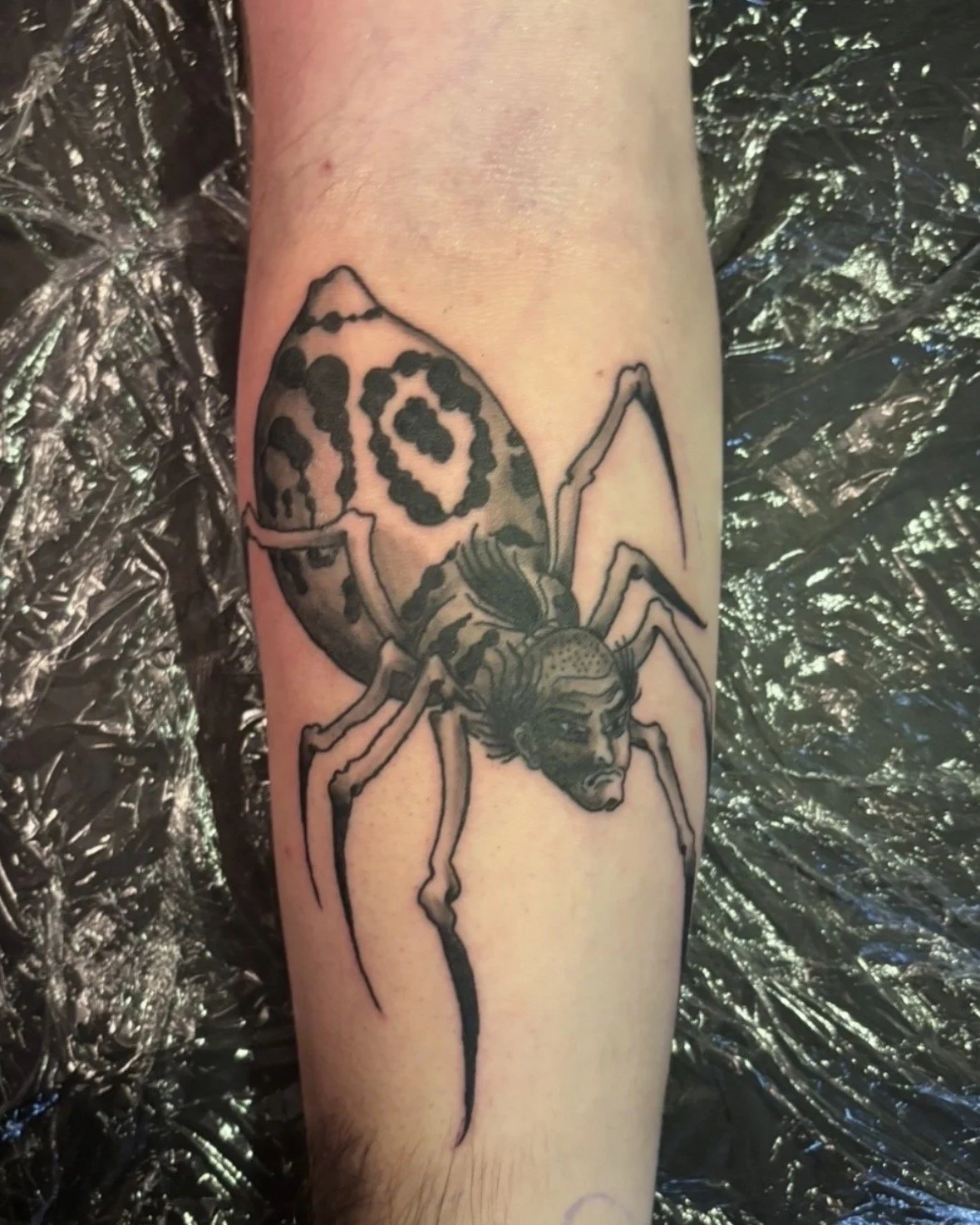 Tattoo of a spider with a human face on its body, featuring black and gray shading, on a person's arm.