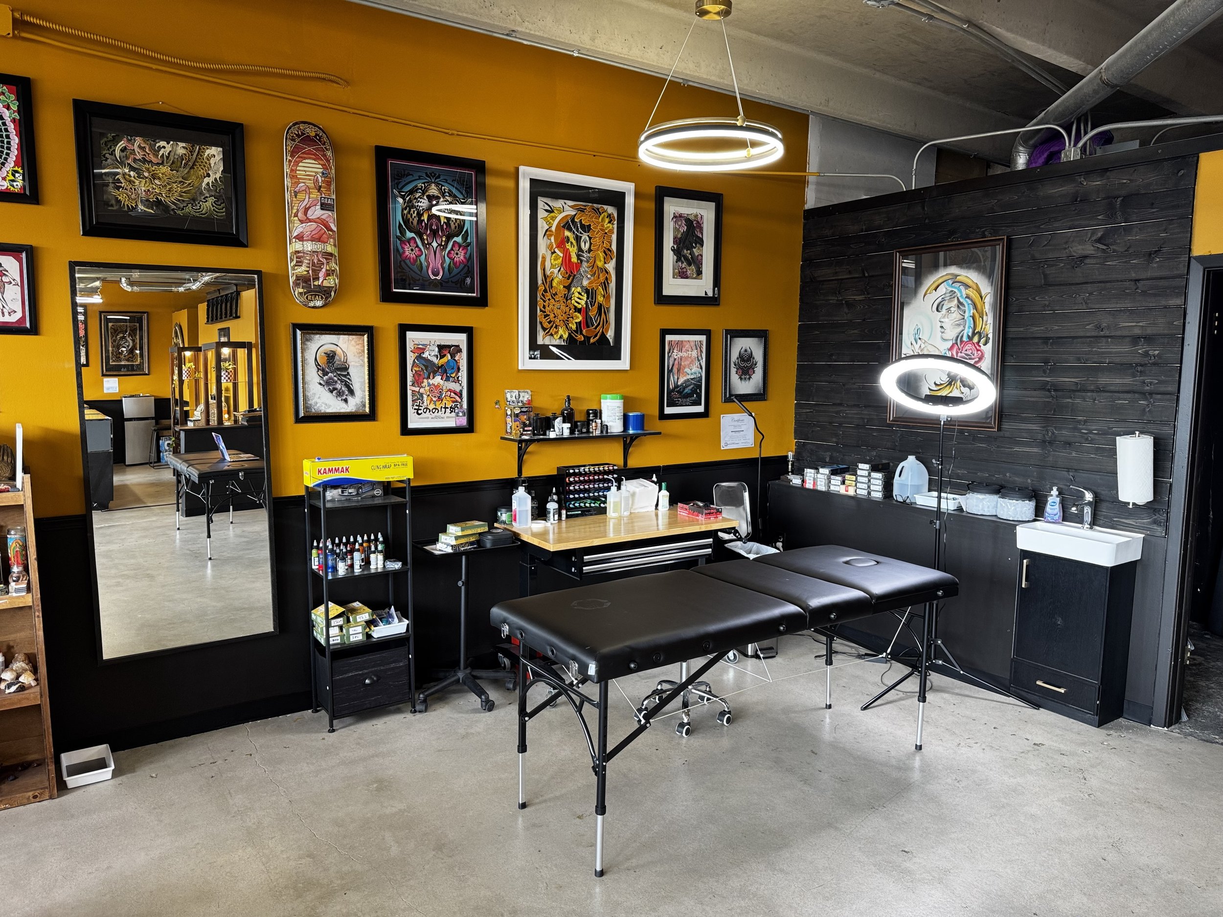 Tattoo shop interior with vibrant artwork on yellow and black walls, tattooing equipment, and a tattoo chair in the foreground.