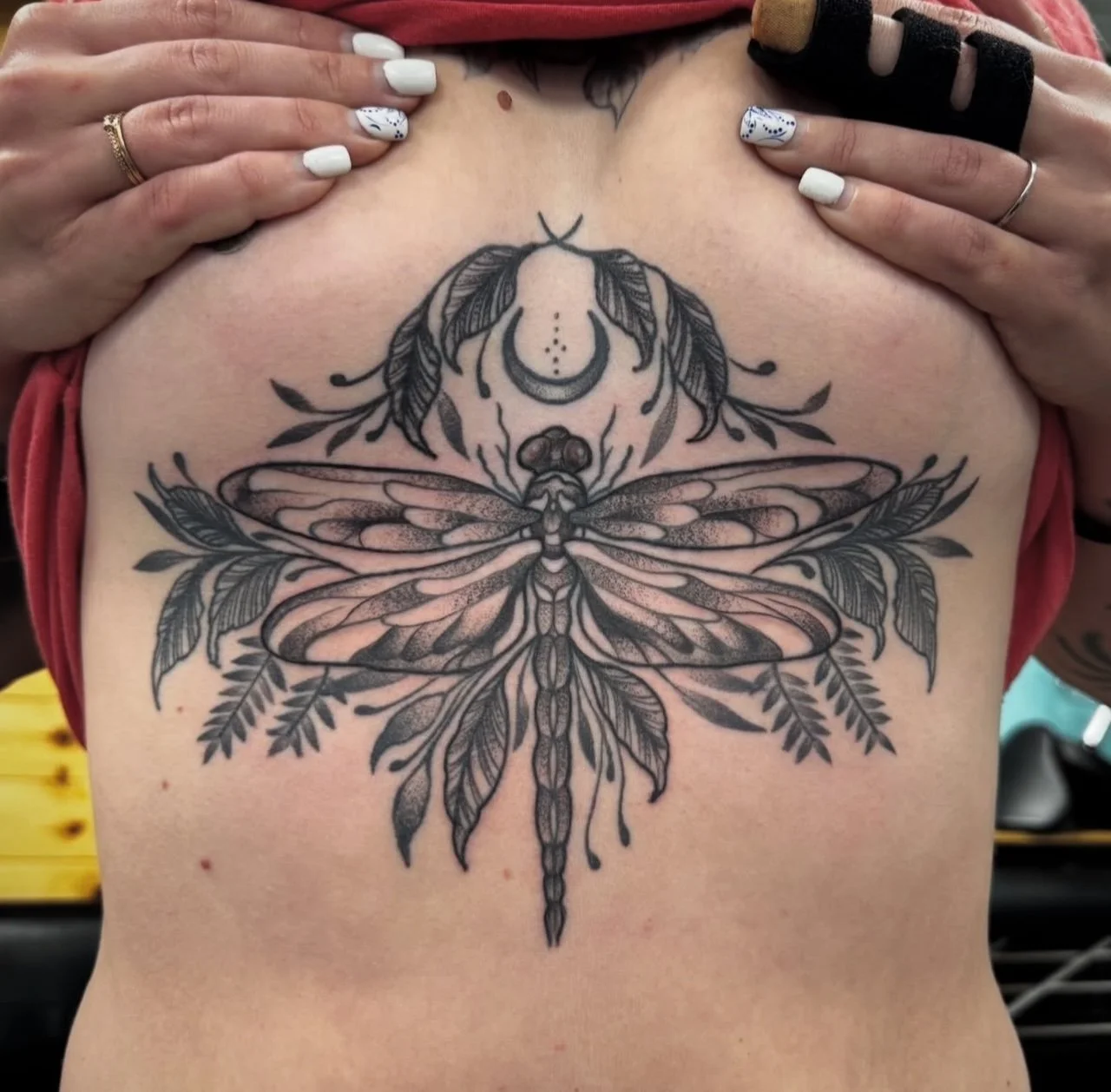 A detailed black and gray tattoo of a dragonfly on a person's chest, finished in the 80033 zip code area.