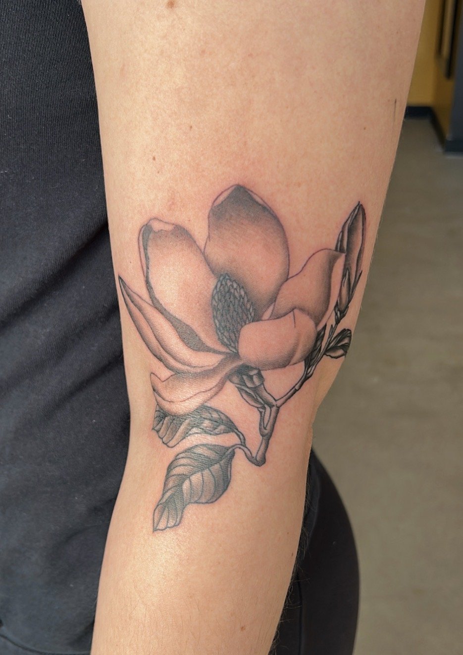 Black and gray tattoo of a magnolia flower with leaves on person's arm completed at our Wheat Ridge shop.