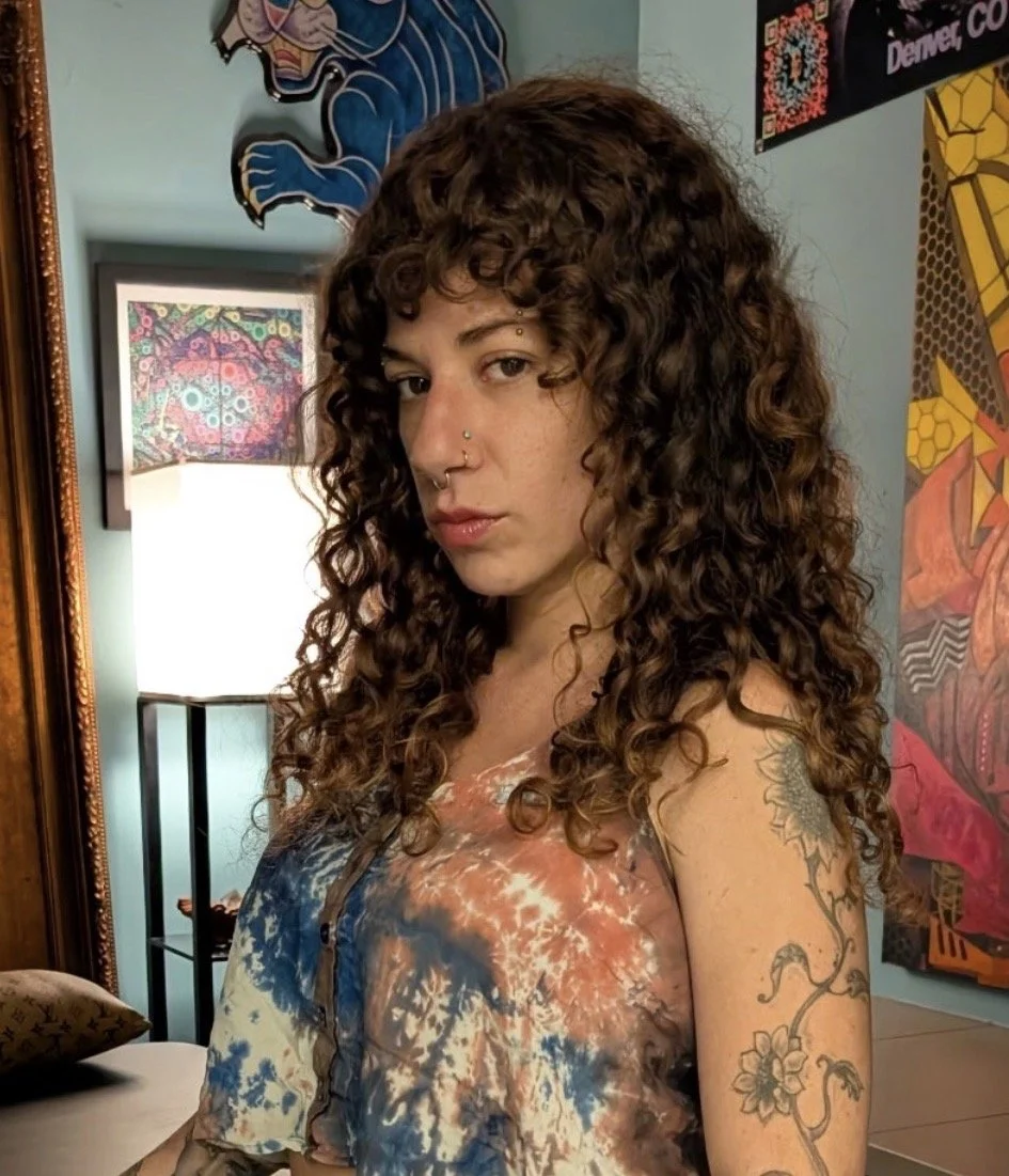Young woman with curly brown hair, nose piercings, and tattoos on her arm, wearing a tie-dye shirt, is standing indoors near a mirror, with a lamp, colorful artwork, and a poster visible in the background.