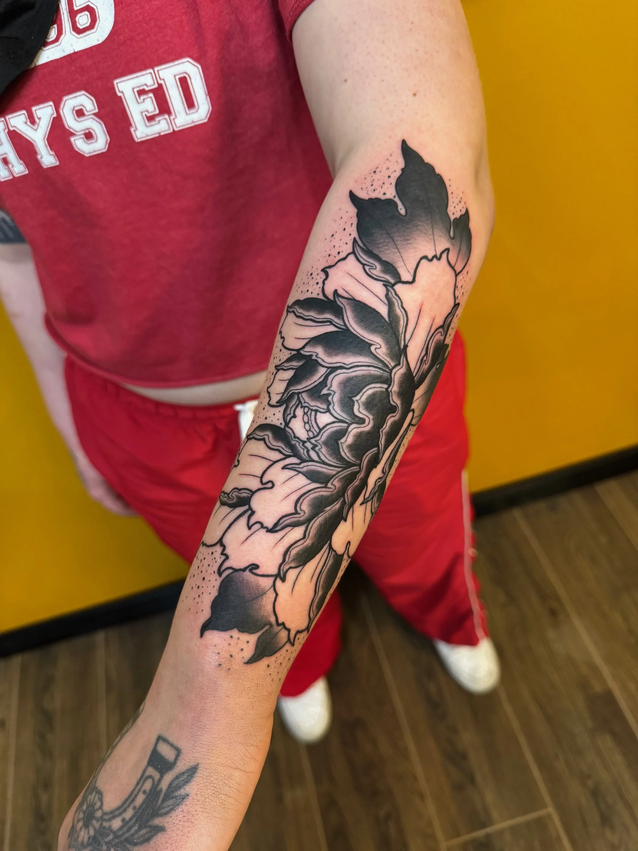Close-up of a person's arm with a black and gray floral tattoo, wearing a red shirt, red shorts, and white sneakers, standing on wooden flooring