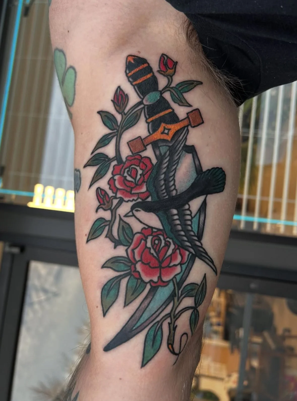 Tattoo of a bird perched on a branch with red roses and green leaves, with a small orange and black butterfly above the flowers.