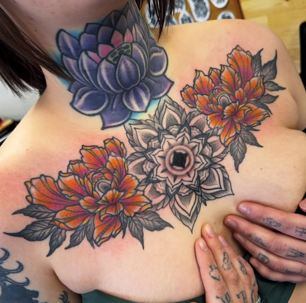 Colorful floral tattoos on a person's chest, including large flowers in shades of purple, orange, and black and white, with the person touching their chest and tattoos on their fingers.