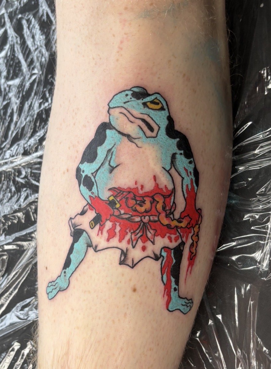A tattoo of a cartoon-style anthropomorphic frog with blue and black skin, a stern expression, and yellow eyes. The frog is dressed in a bloodied white karate gi with a red belt and is holding nunchucks that are also covered in blood.