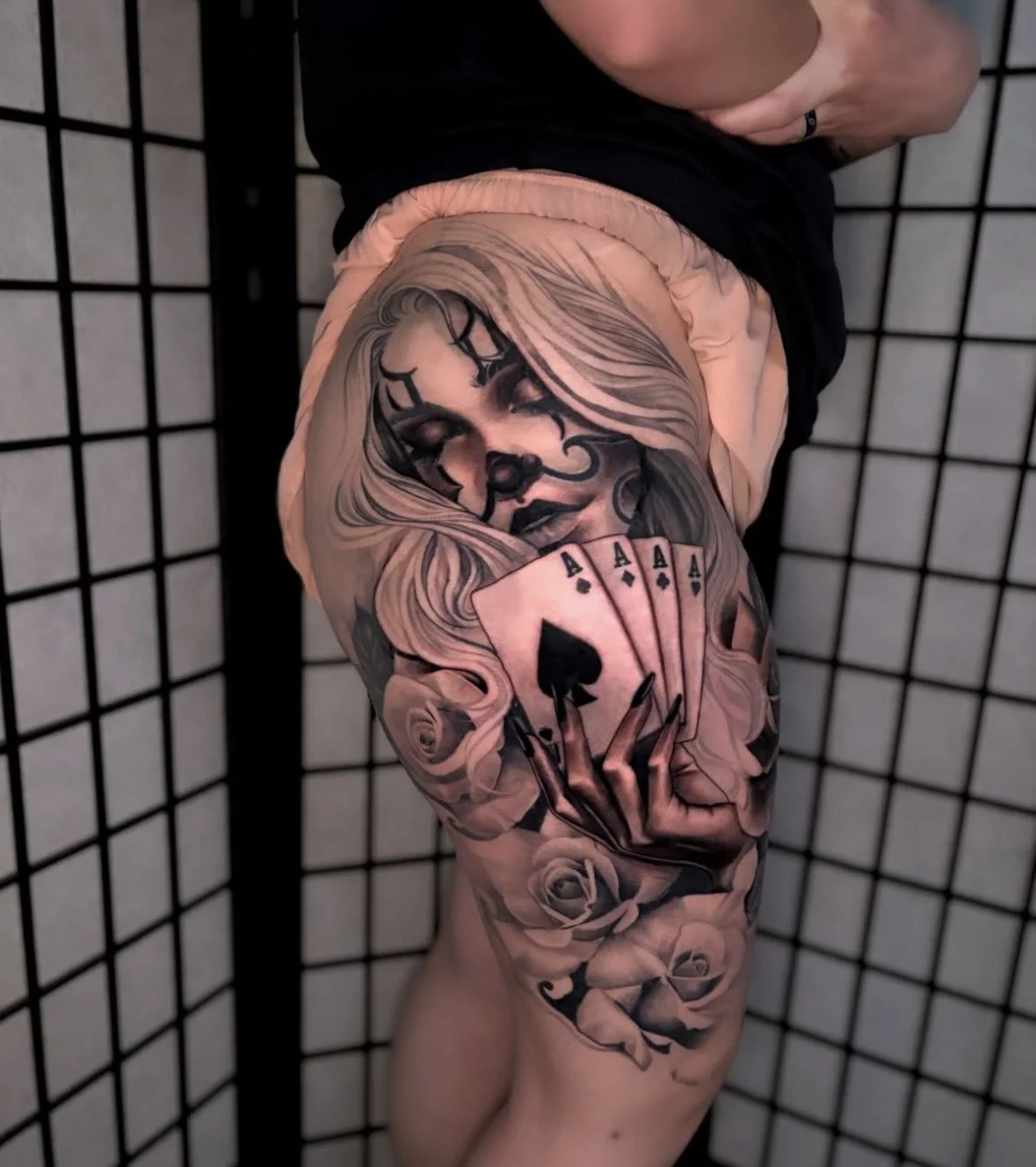 Tattoo of a woman with dark makeup and flowing hair playing cards and holding 4 aces, surrounded by roses, located on the upper thigh.