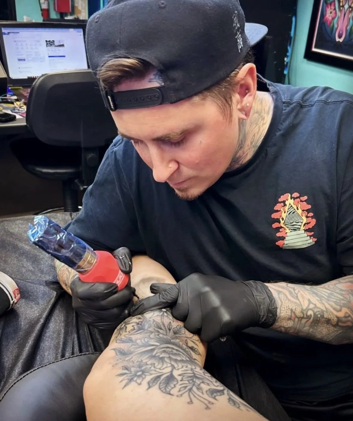 A tattoo artist with tattoos on his arms, wearing a black baseball cap backward and black gloves, tattooing a person's arm in a tattoo studio.