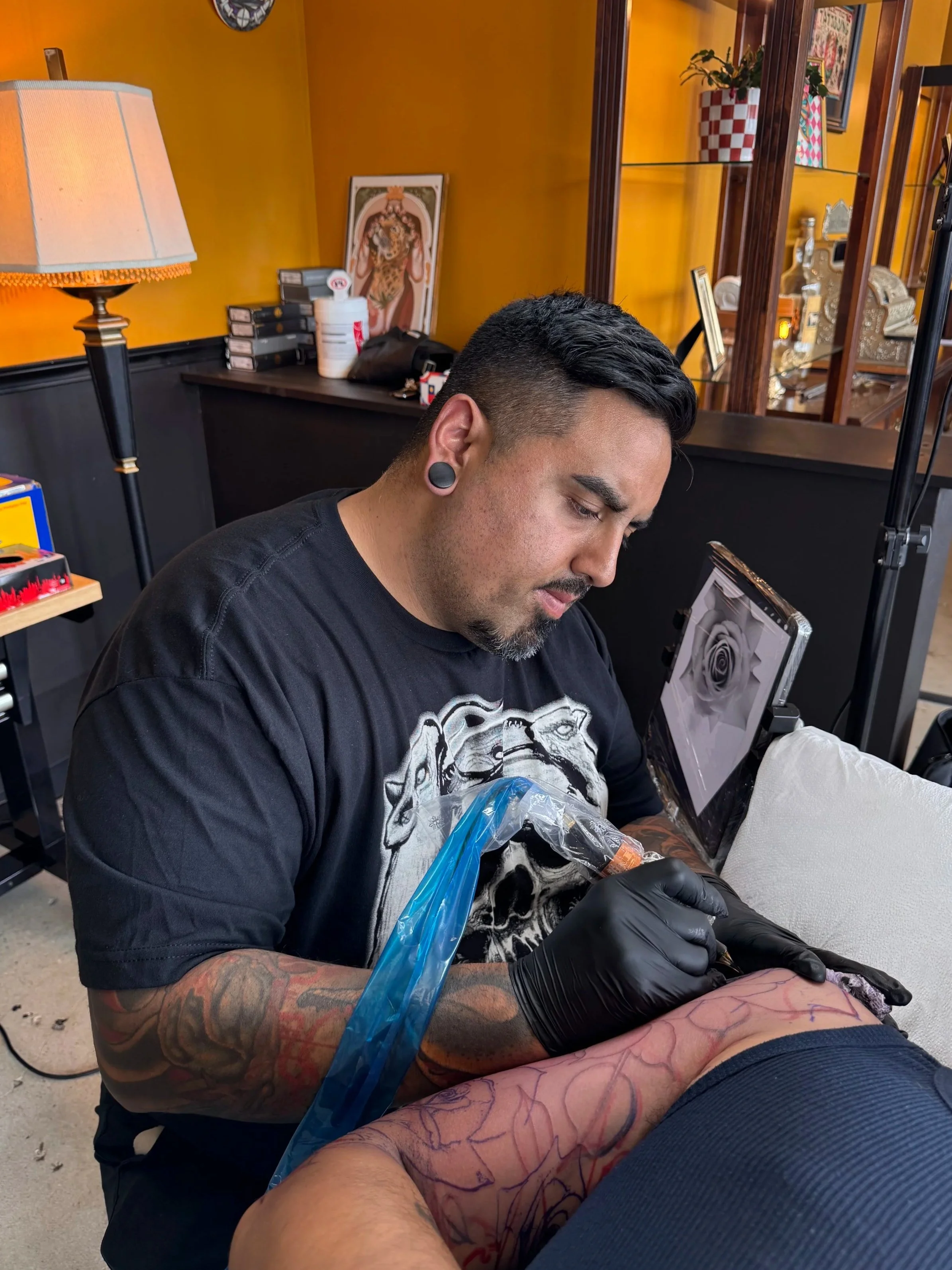 A tattoo artist with short dark hair, tattoos on his arms, black gloves, and large gauge earrings, is tattooing a person's leg in a room with yellow walls, a lamp, and various decorative items and artwork on shelves.