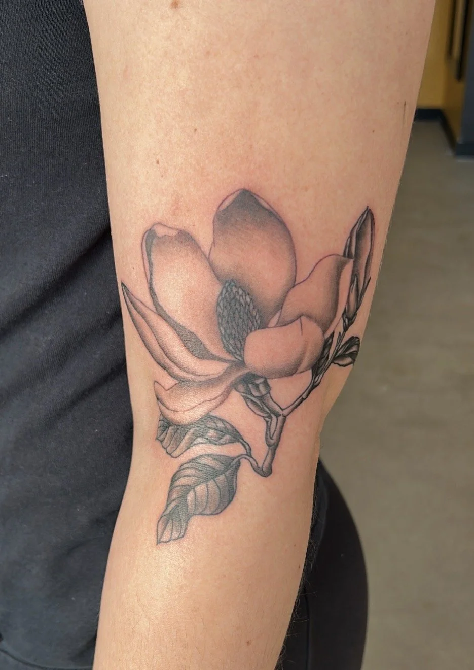 A black and gray line art tattoo of a magnolia flower on a person's forearm.