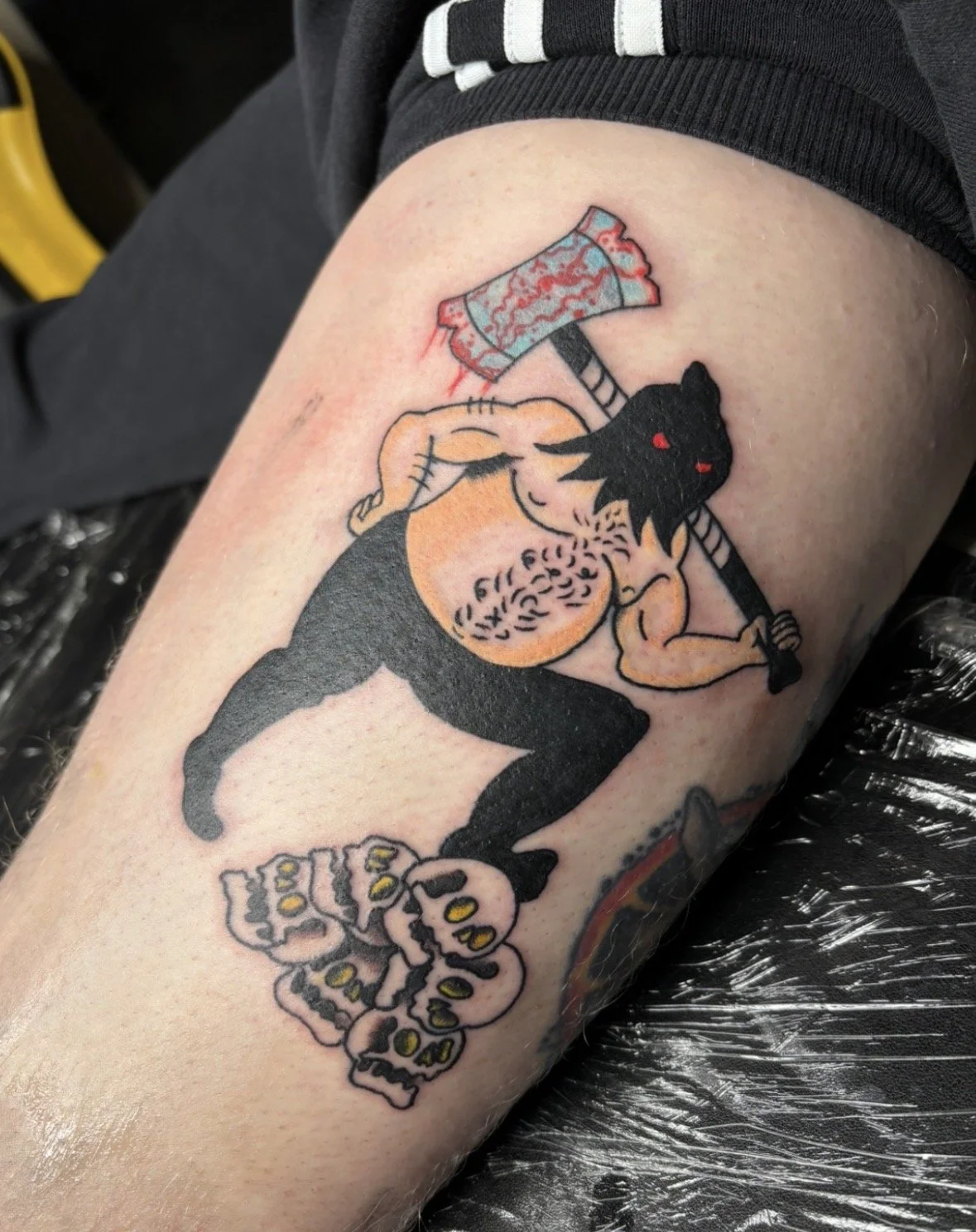 Tattoo of a muscular shirtless man with a black mask and red eyes, holding a bloody axe, and a skull at his feet.
