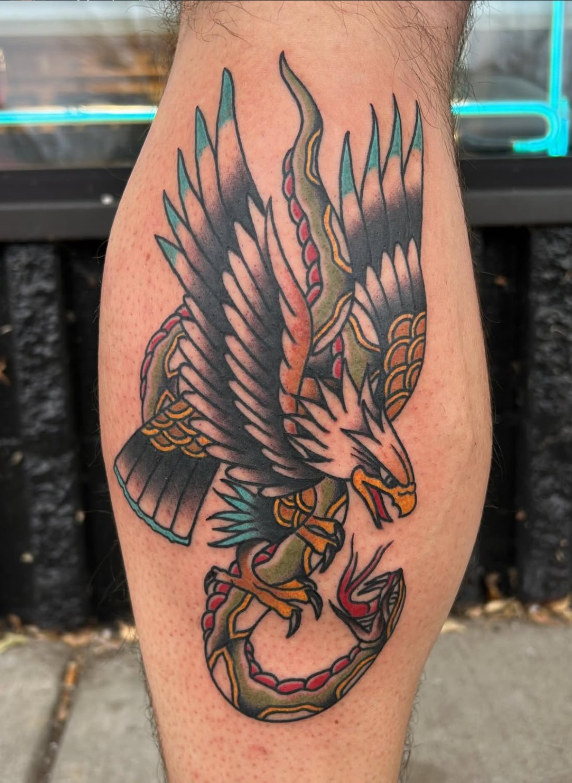 A traditional tattoo of a stylized eagle with wings spread, holding a snake in its talons, with a fierce expression.