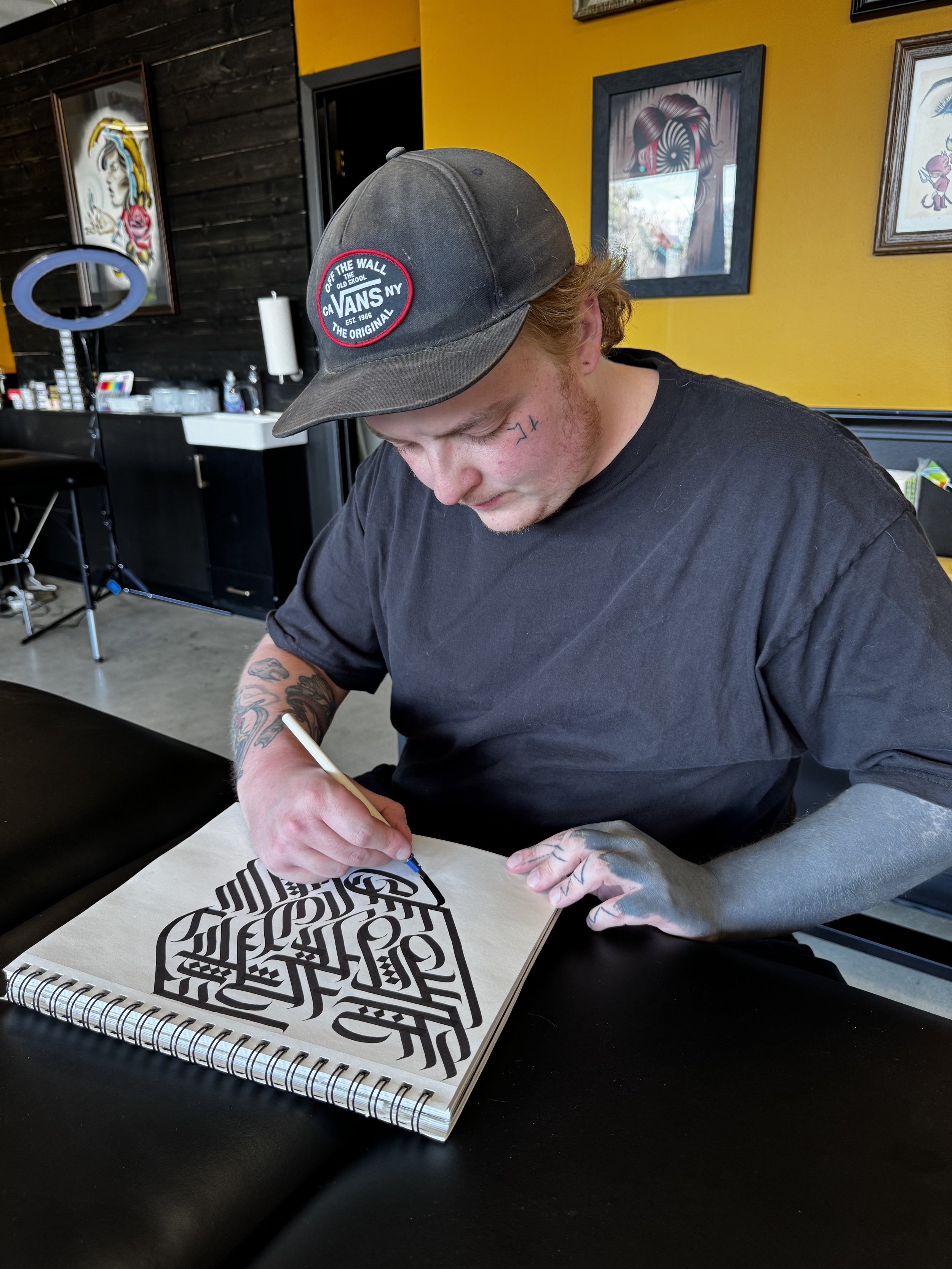 A young man with tattoos on his arms, wearing a black T-shirt and a dark baseball cap with a logo, is drawing intricate black and white geometric art in a sketchbook.