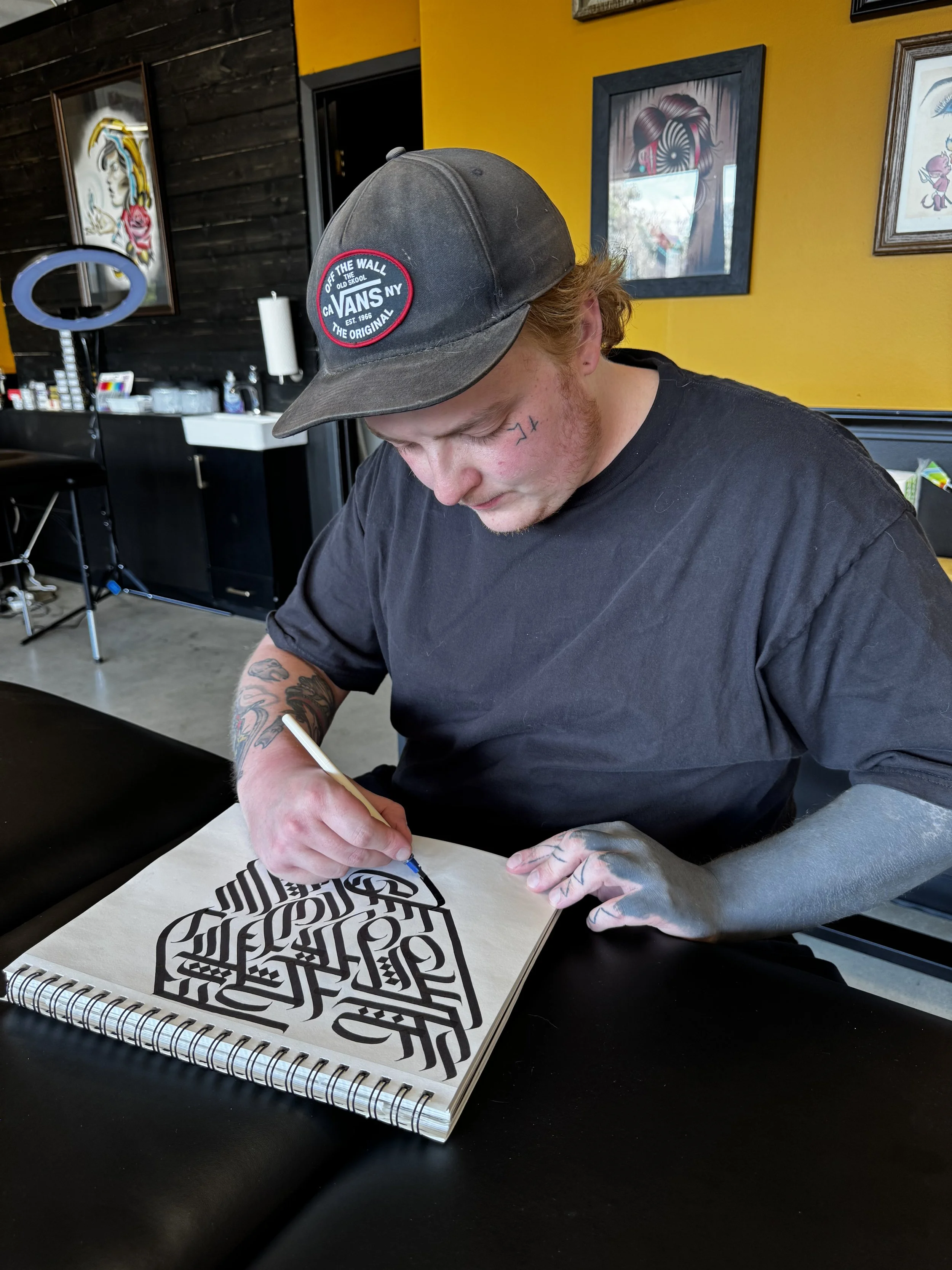 A young man with tattoos on his arms, wearing a black T-shirt and a dark baseball cap with a logo, is drawing intricate black and white geometric art in a sketchbook.