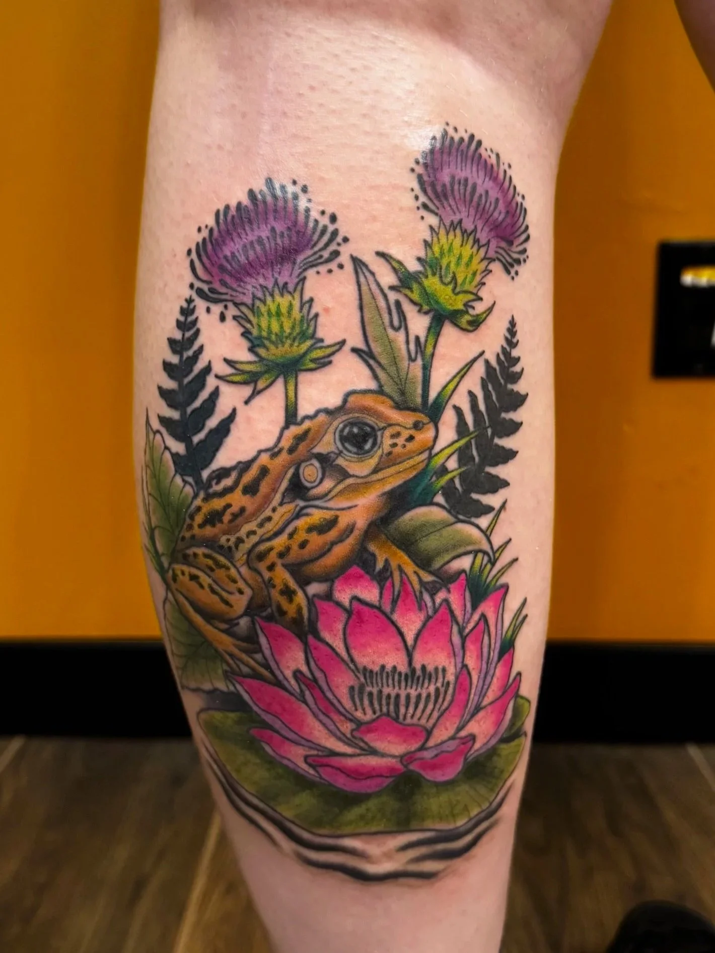 Colorful tattoo of a yellow frog among pink lotus flowers, green leaves, and other purple flowers on a person's calf.