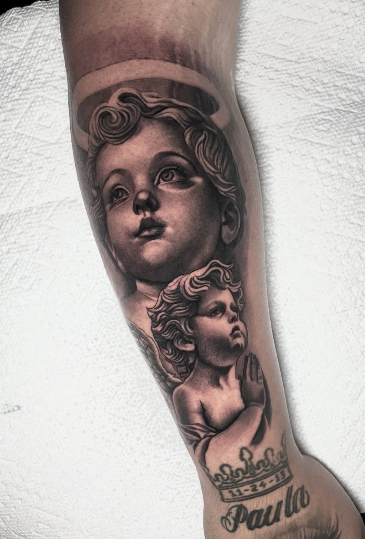 Tattoo of angelic figures on a person's forearm, with a date 11-24-13 and the name Paula below.