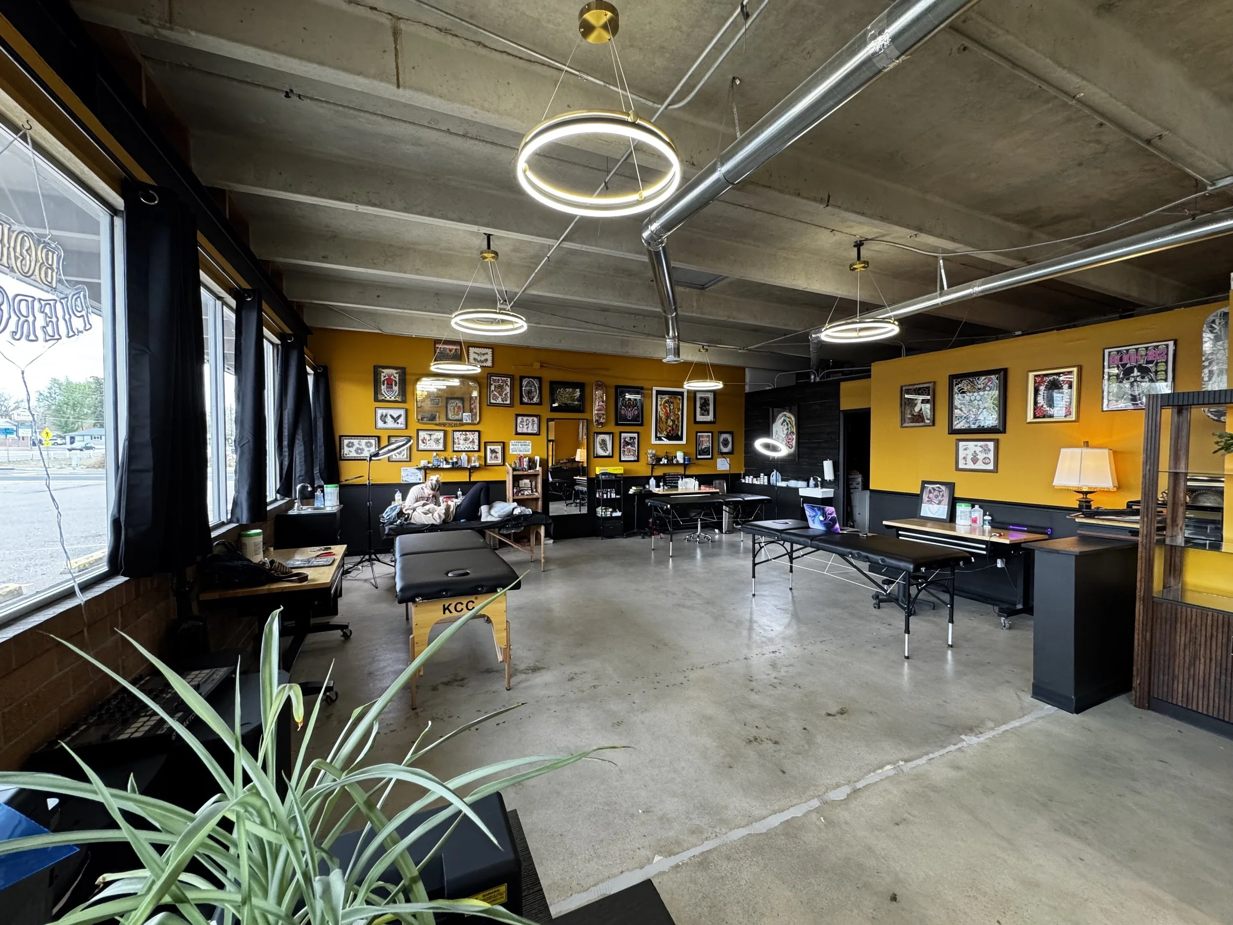 Interior of a modern tattoo studio with exposed concrete ceiling, large windows with black curtains, and yellow walls decorated with framed tattoo artwork. There are several black benches and tables, a person lying on a black chair, and bright circular ceiling lights.